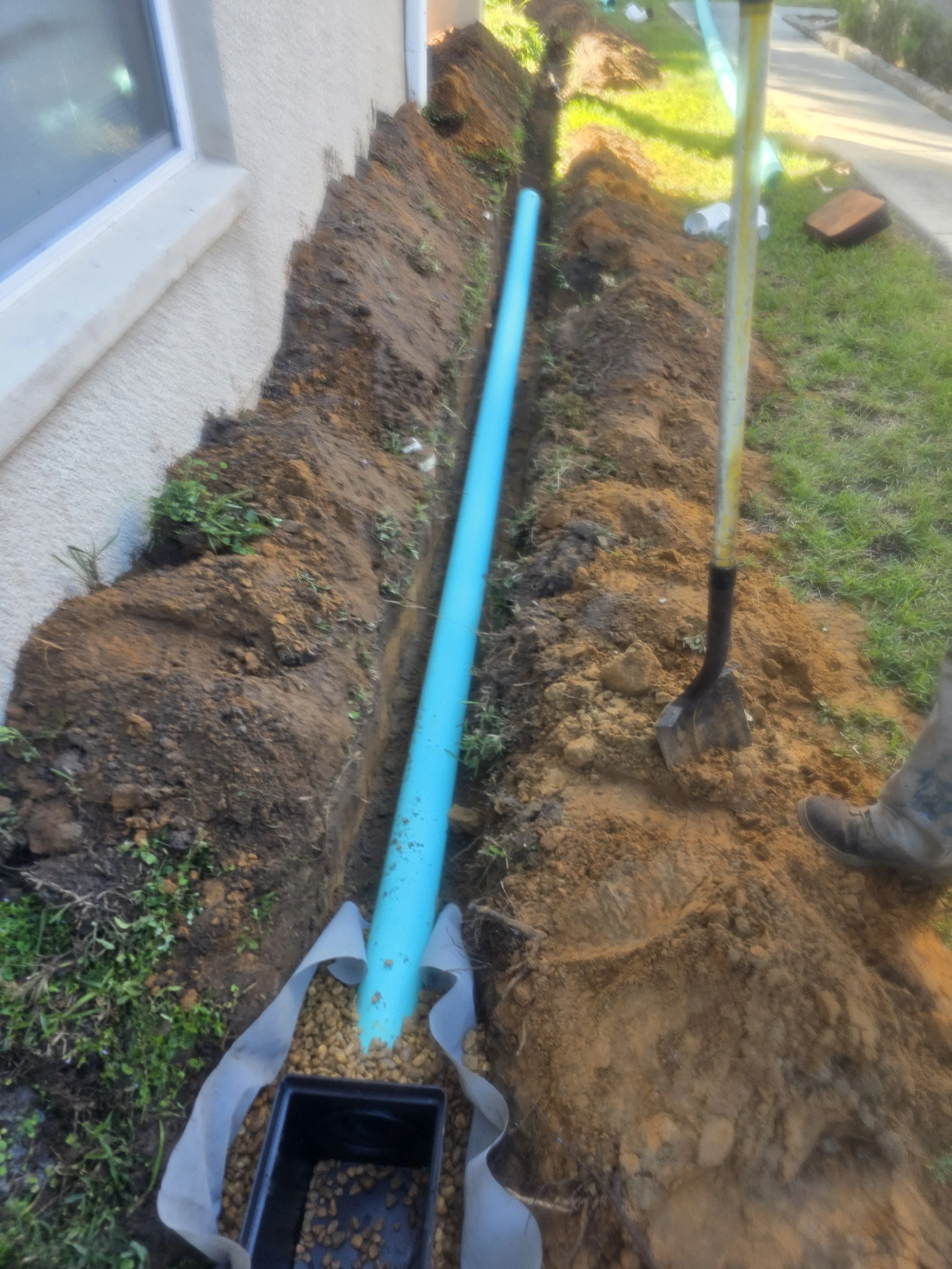 A trench dug along the side of a house with a blue pipe installed inside, and construction tools such as a shovel present nearby, in a grassy yard.