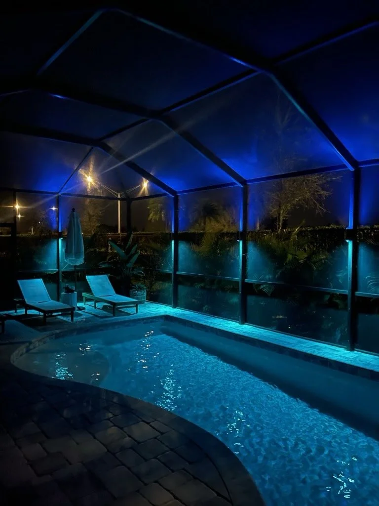Nighttime view of a small pool enclosed by a screened structure with blue and warm yellow lighting, poolside lounge chairs, a closed umbrella, and lush greenery outside.