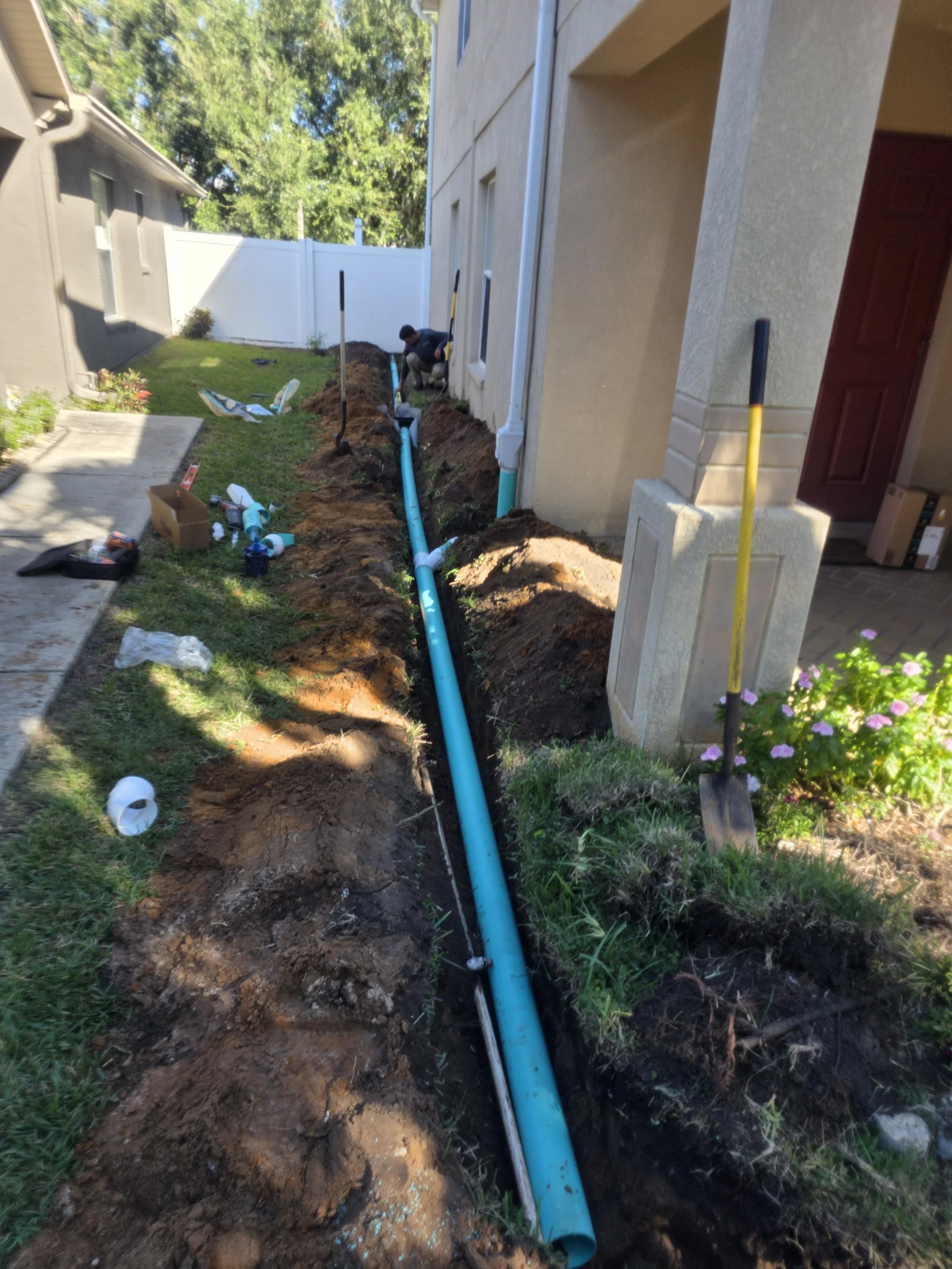 Workers installing a blue underground pipe in a trench beside a residential house.