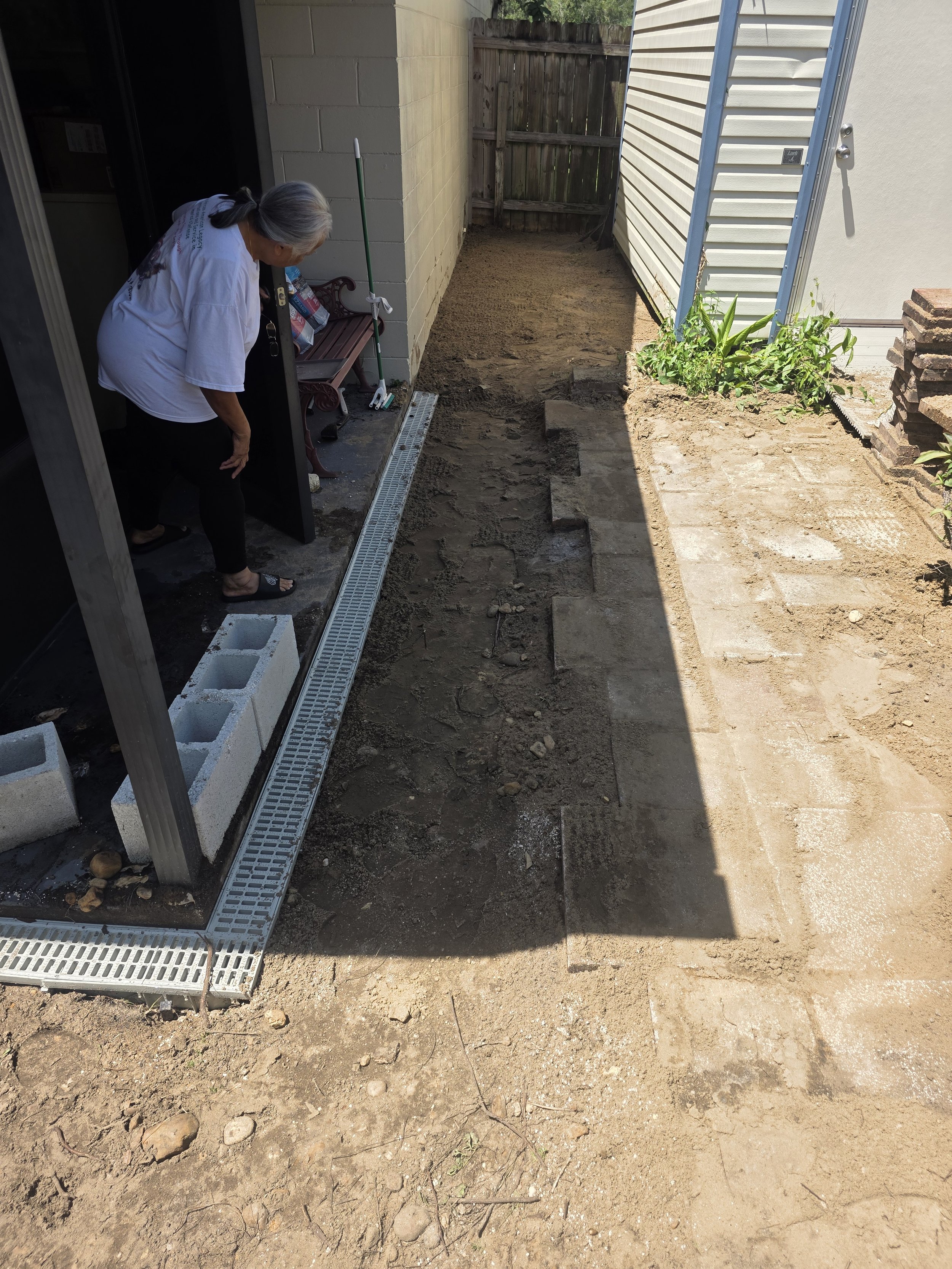 A person working on a small backyard landscaping project, laying down bricks for a patio next to a house with a white exterior and a wooden fence behind.