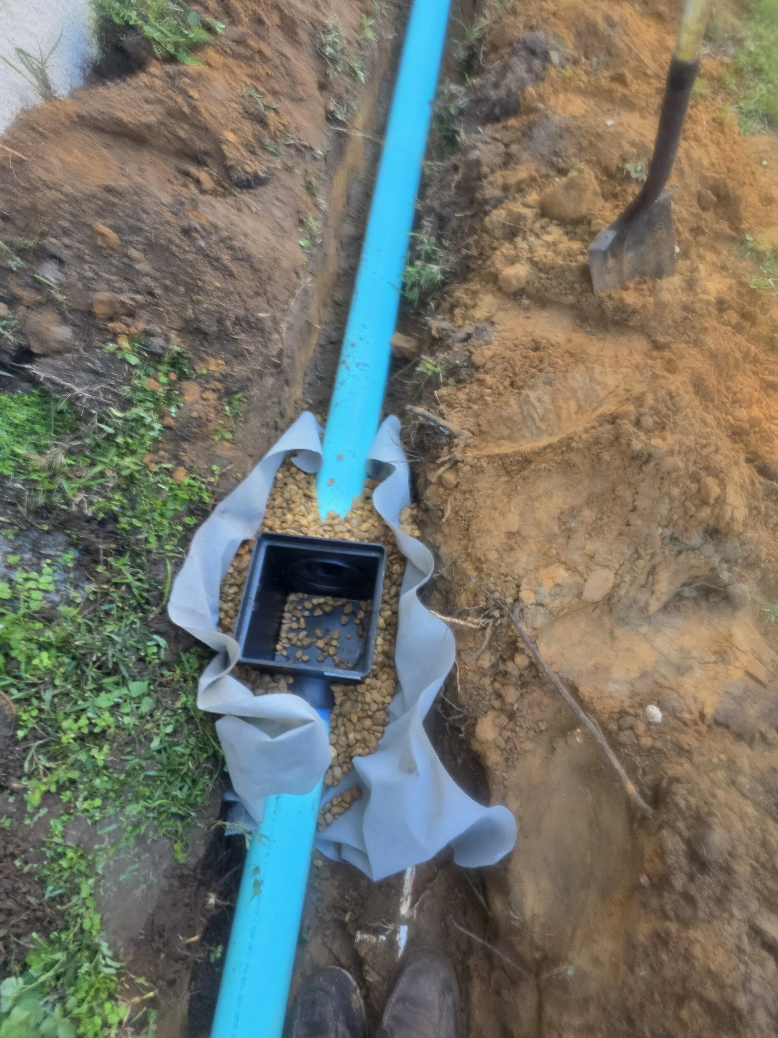 Installing a blue plastic pipe in a trench, lined with a fabric filter, with gravel in the pipe and a shovel nearby on the dirt ground.