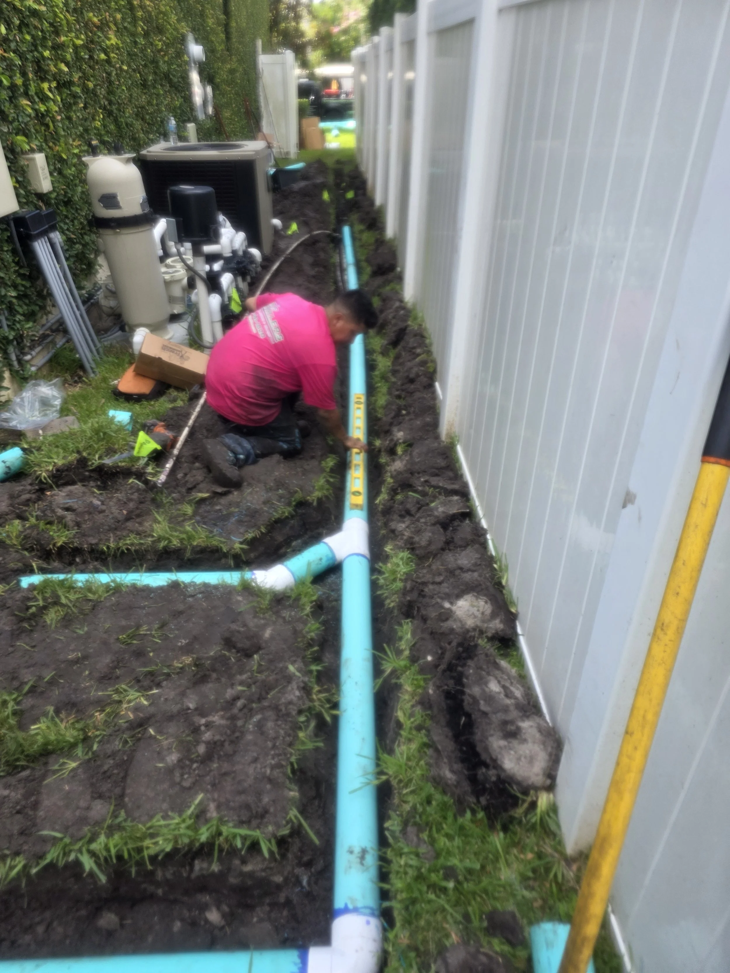 A person working on installing or repairing blue underground pipes along a white fence in a backyard. The person is kneeling on the ground, surrounded by tools and equipment, with a passageway filled with dirt and small plants.
