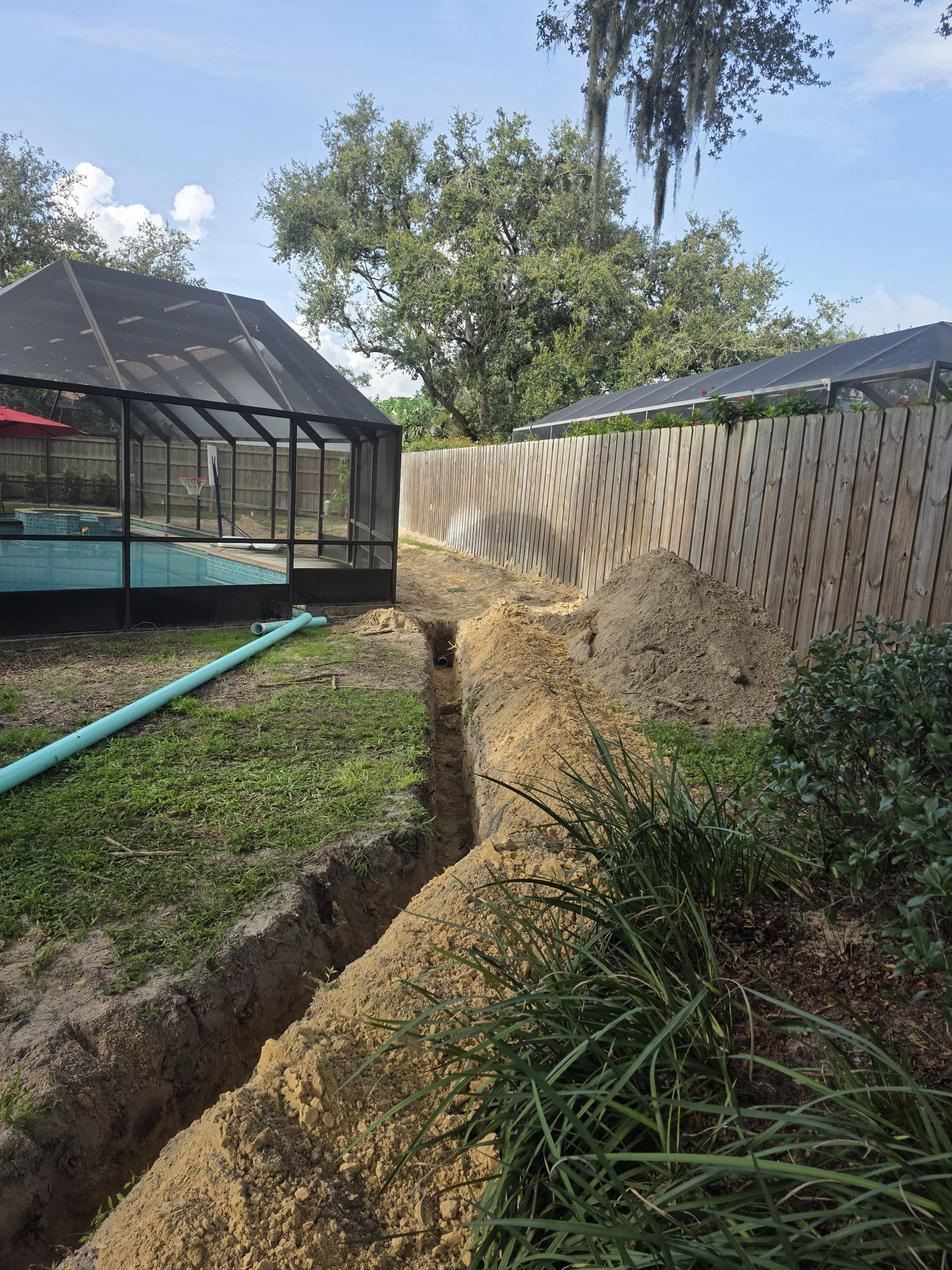 A backyard with an in-ground swimming pool, a screened enclosure, and a wooden fence. There is a trench being dug with a pile of dirt nearby, and gardening plants along the edge.