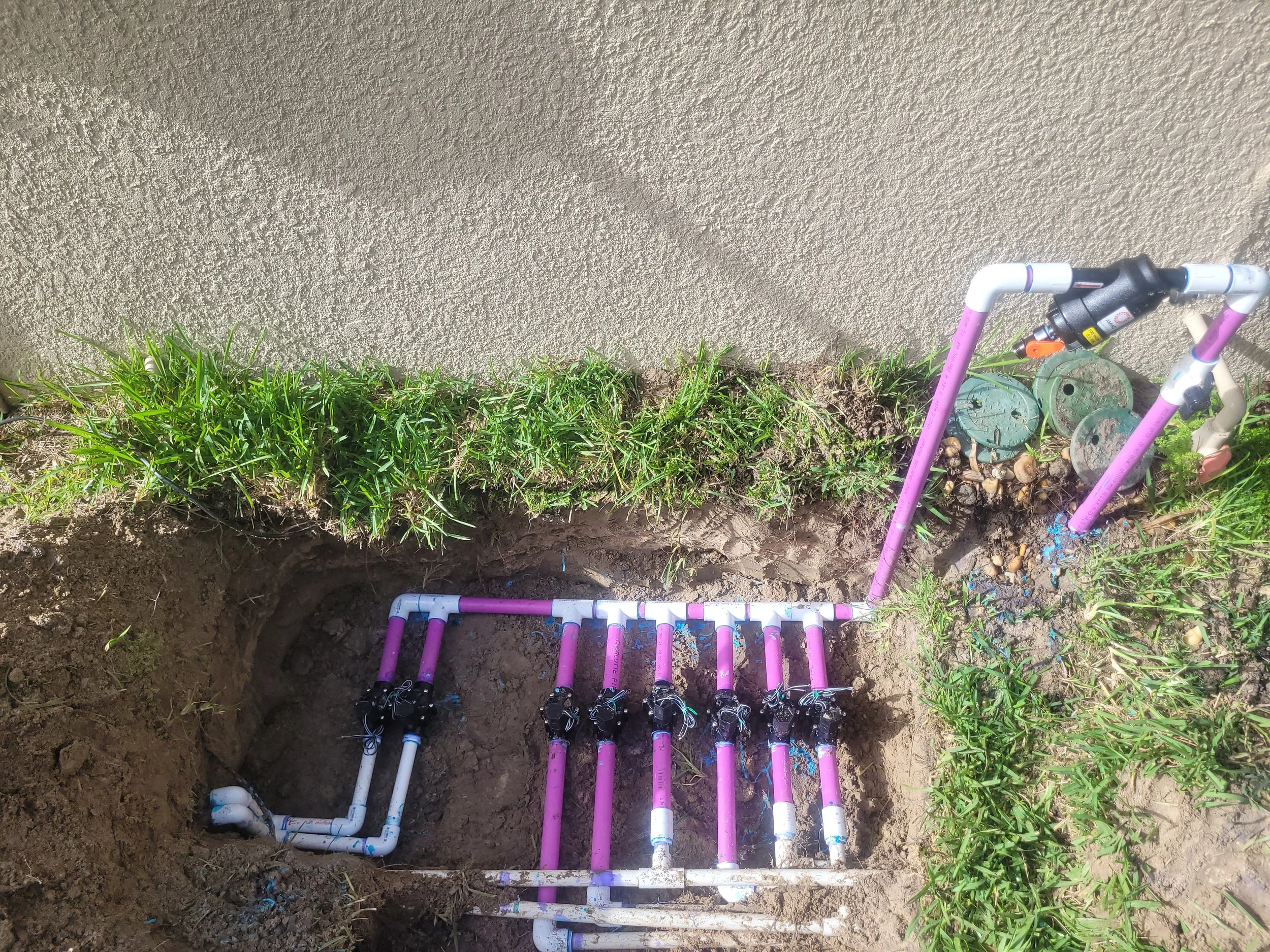 A plumbing installation in a trench featuring pink and white PVC pipes connected with black fittings, with a valve at the top right, surrounded by soil, grass, and some green utility covers.