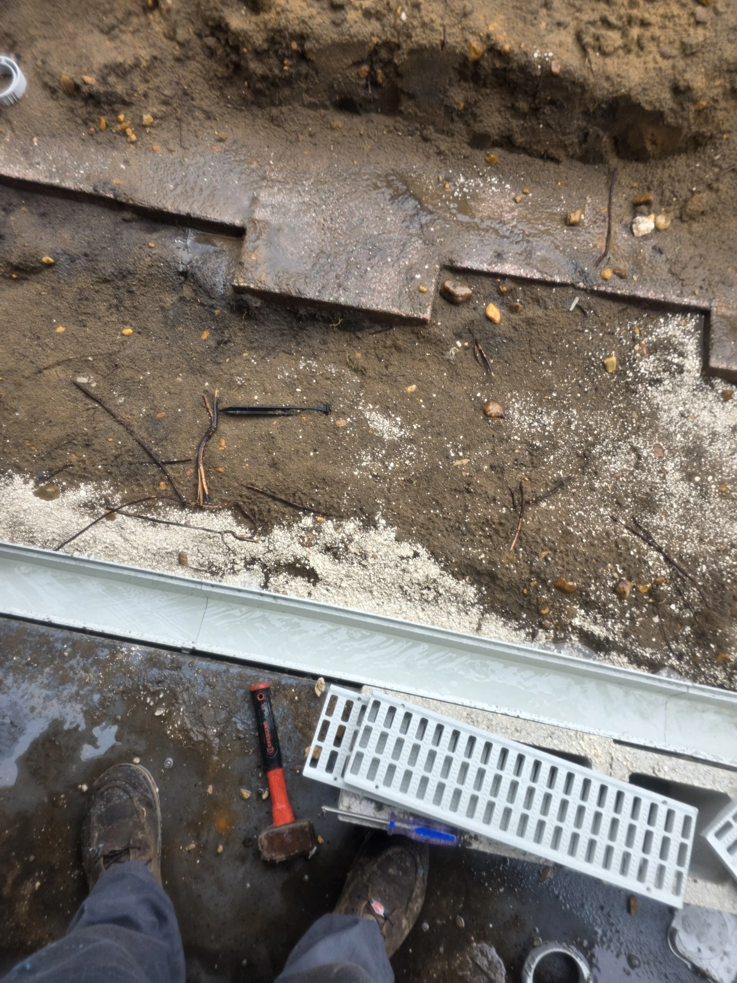 Construction worker floor area with tools, including a hammer and a drill, next to a metal grate and a drainage area, with exposed soil and concrete.