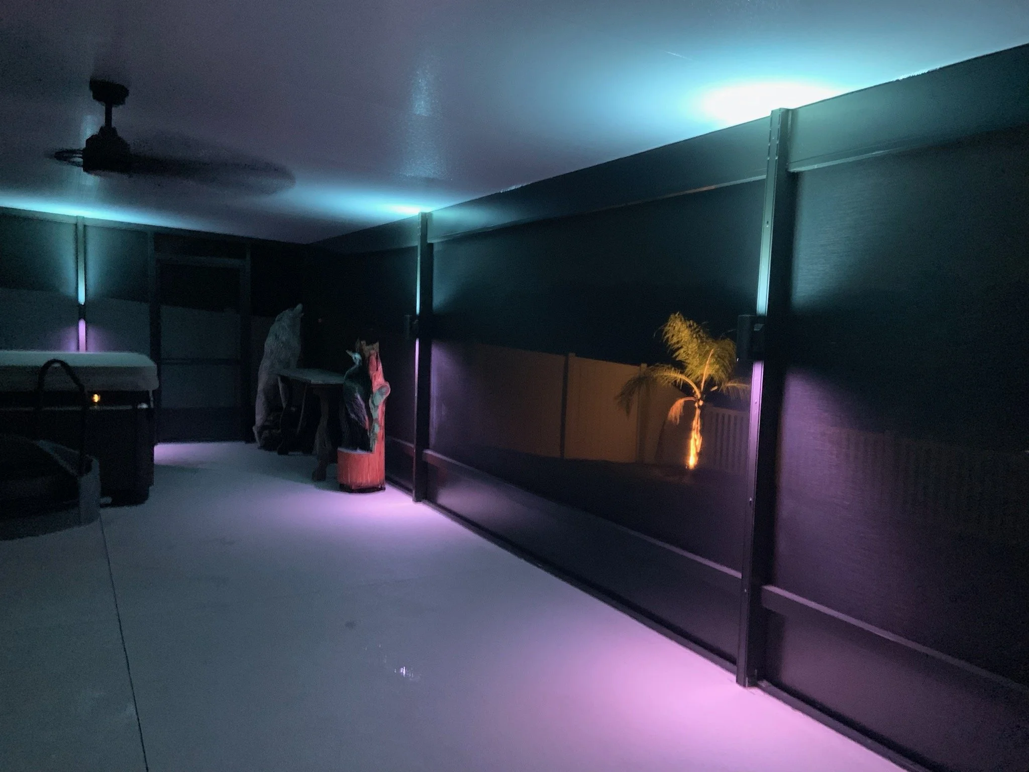 An indoor room with a ceiling fan, various clothing items on a stand, a small table, and a large window or balcony door backlit with multicolored LED lights illuminating a small potted palm tree outside.