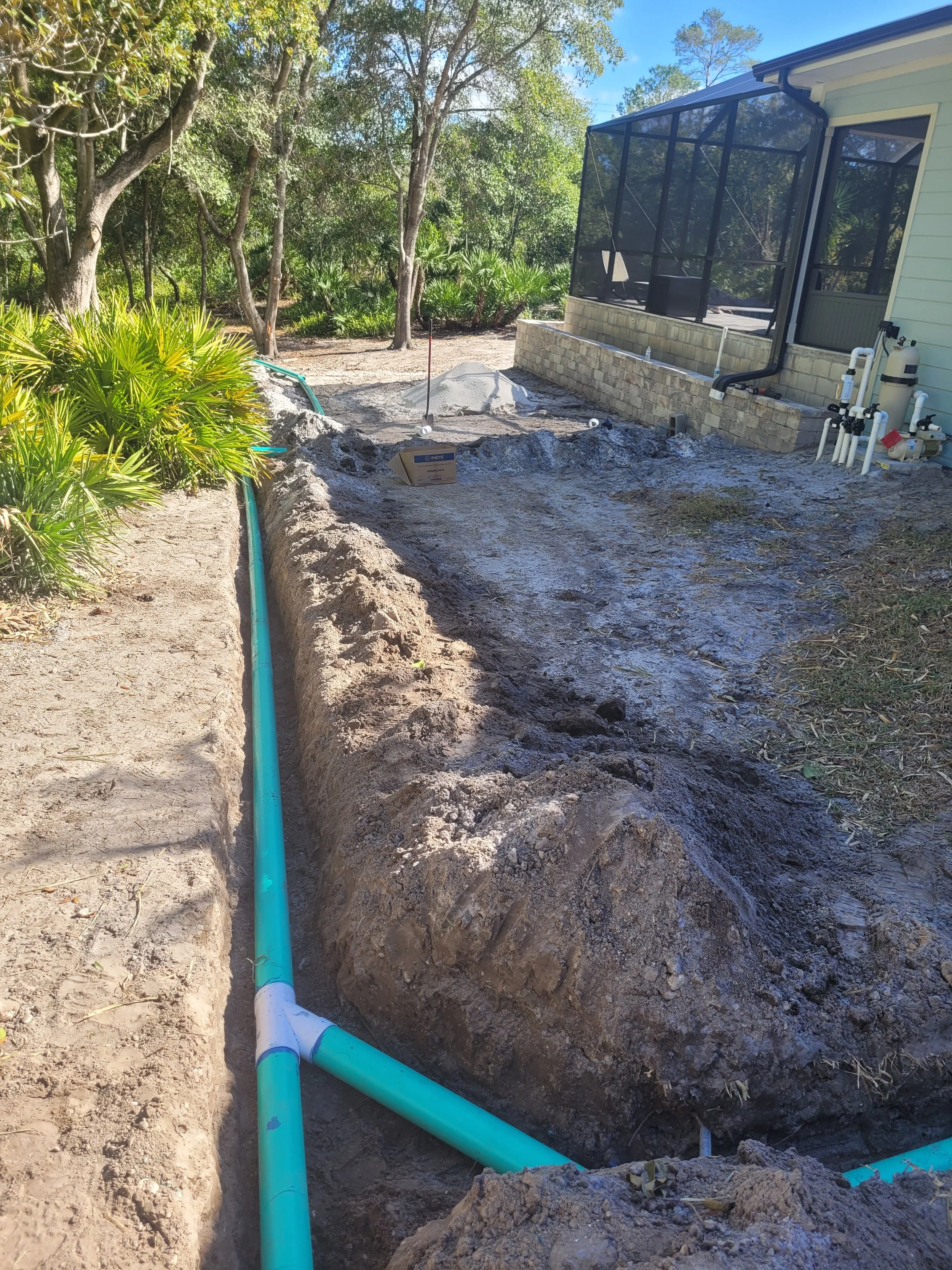 Under construction site showing a trench with blue pipes, a card box, and a pile of soil, next to a house with a screened porch and backyard trees.