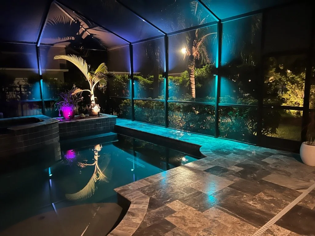 Nighttime view of an enclosed pool area with tropical plants illuminated by colorful lights, including a pink-lit potted plant and palm trees, with a tiled deck surrounding the pool and a glass screen showing outdoor trees and bushes.