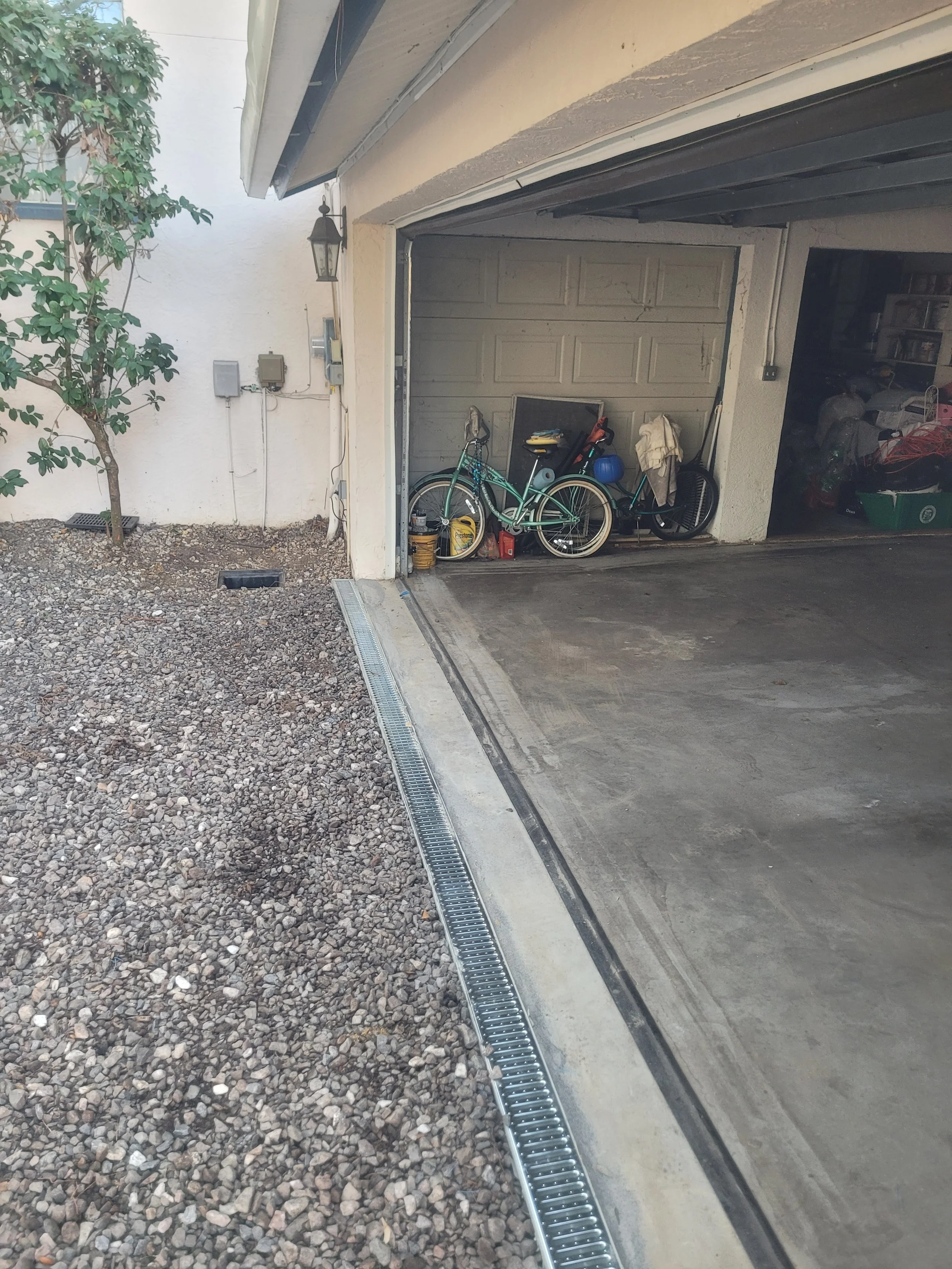 An open garage with bicycles, tools, and storage items inside. Outside, there's a gravel area with a drain and a small tree against a white wall.
