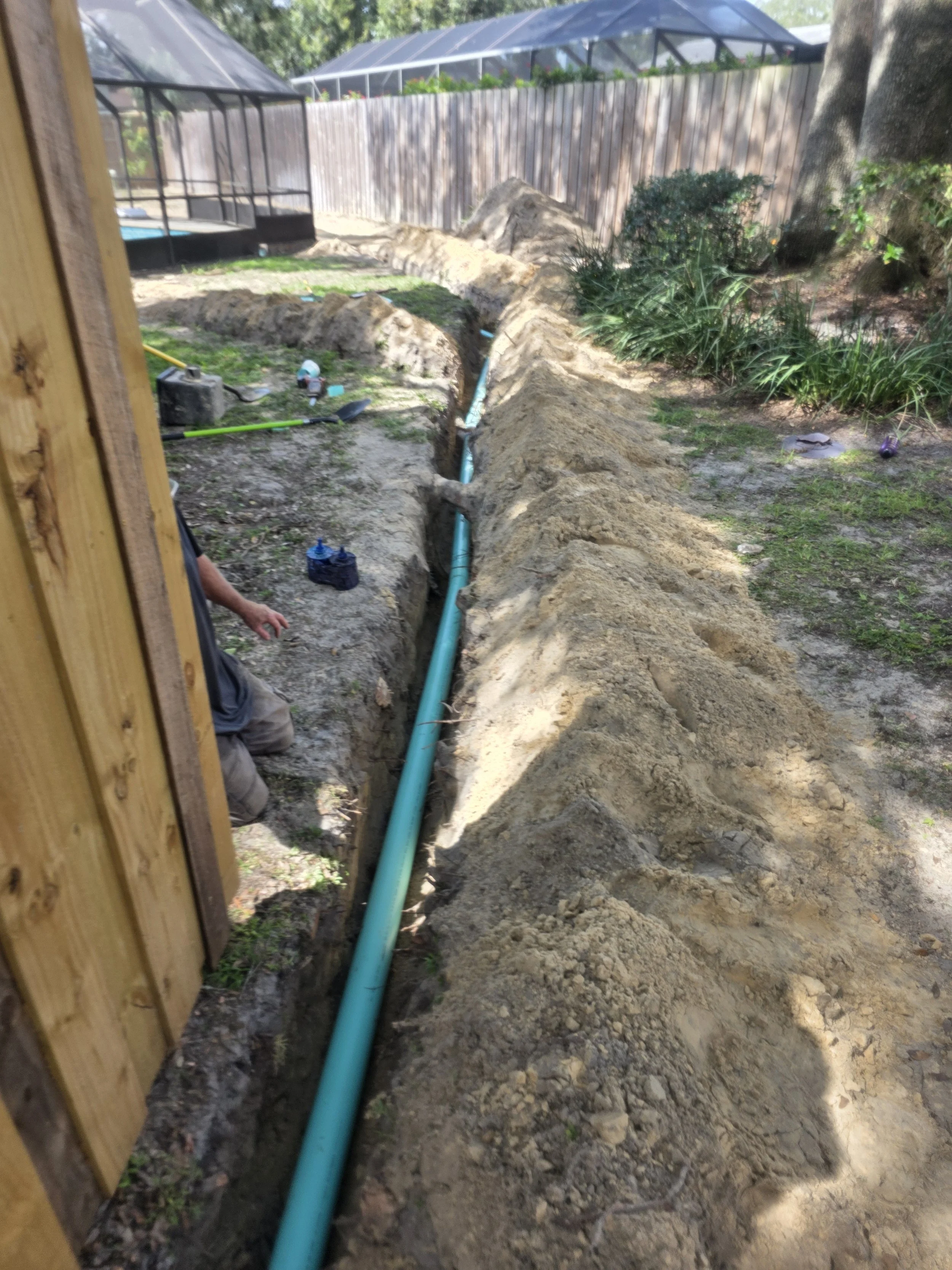 A construction worker is installing a blue pipe in a trench dug in a backyard, with tools and dirt around.