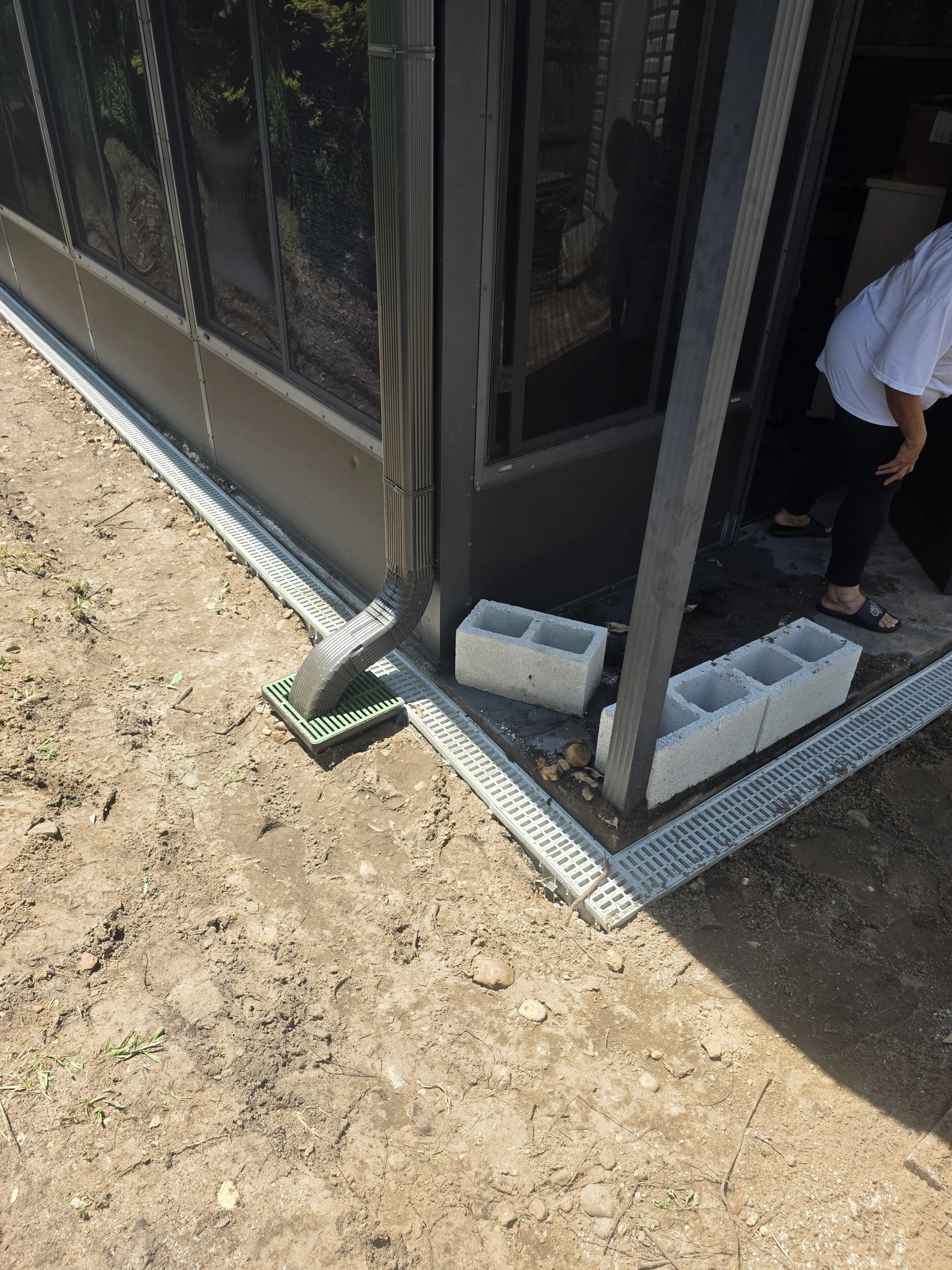 Exterior of a building with a drainage system along the foundation and a downspout directing water into a drain. Cinder blocks are placed nearby.