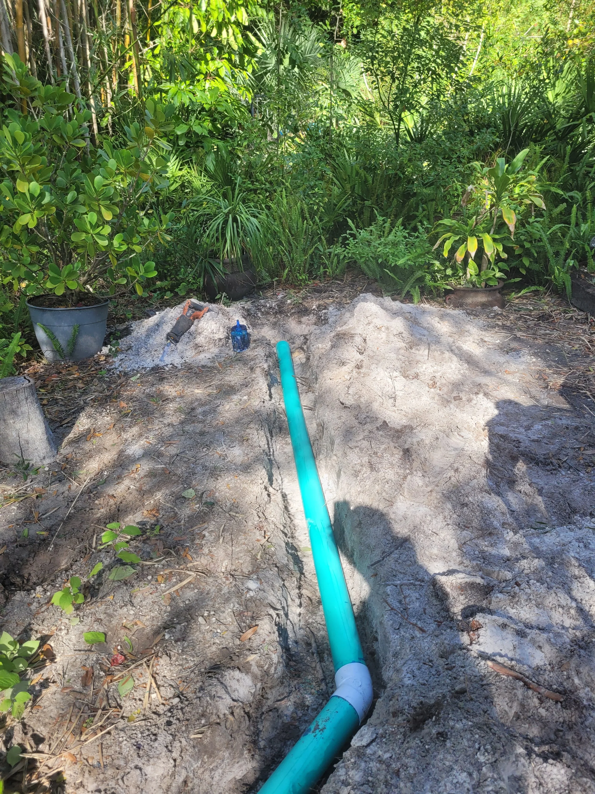 A construction site outdoors with an exposed blue PVC pipe in the ground, surrounded by dirt, with lush green plants and potted plants in the background.