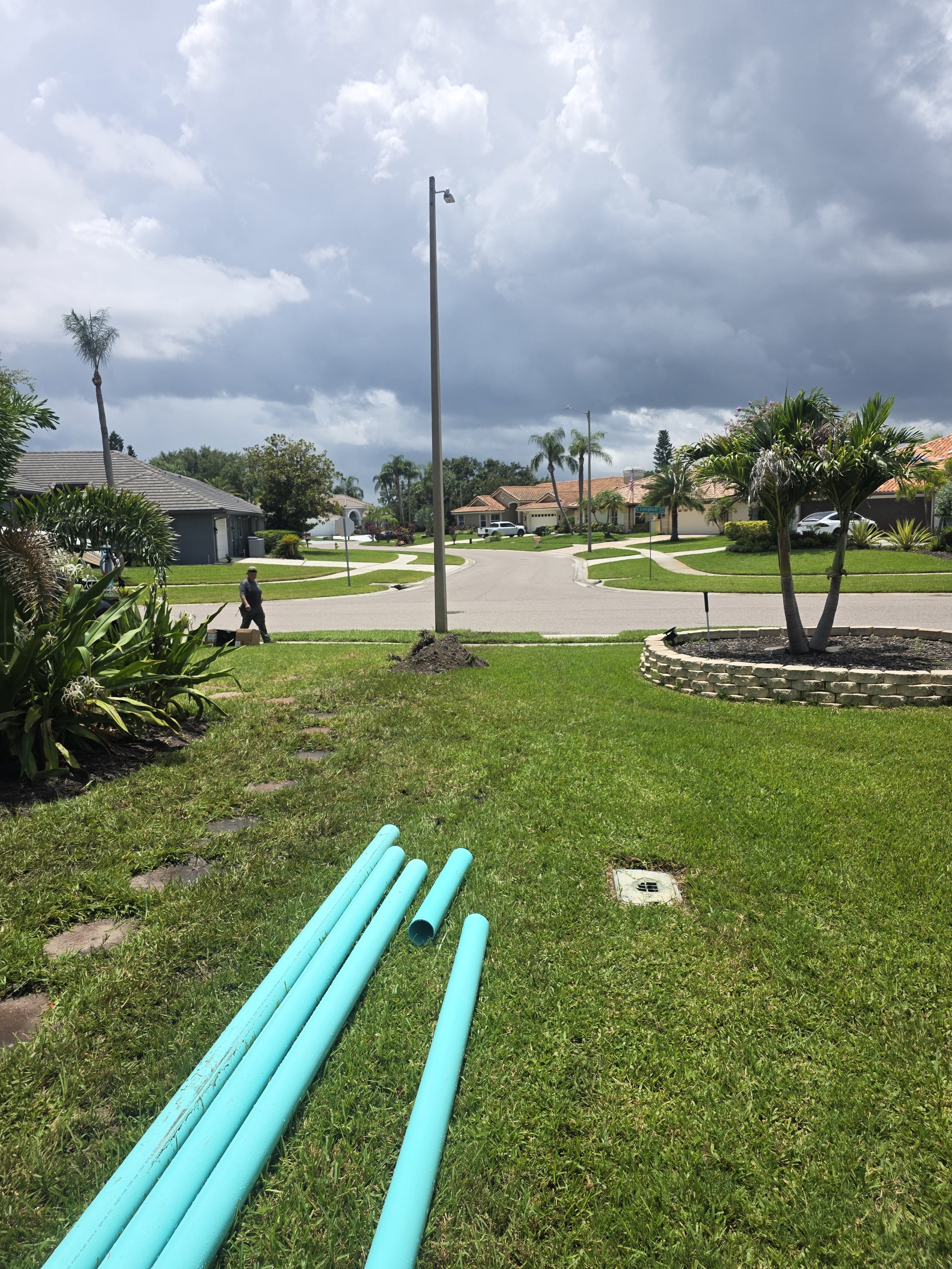 Residential neighborhood street with palm trees, houses, a man walking, and a cloudy sky. In the foreground, green construction pipes are laid on well-kept grass.