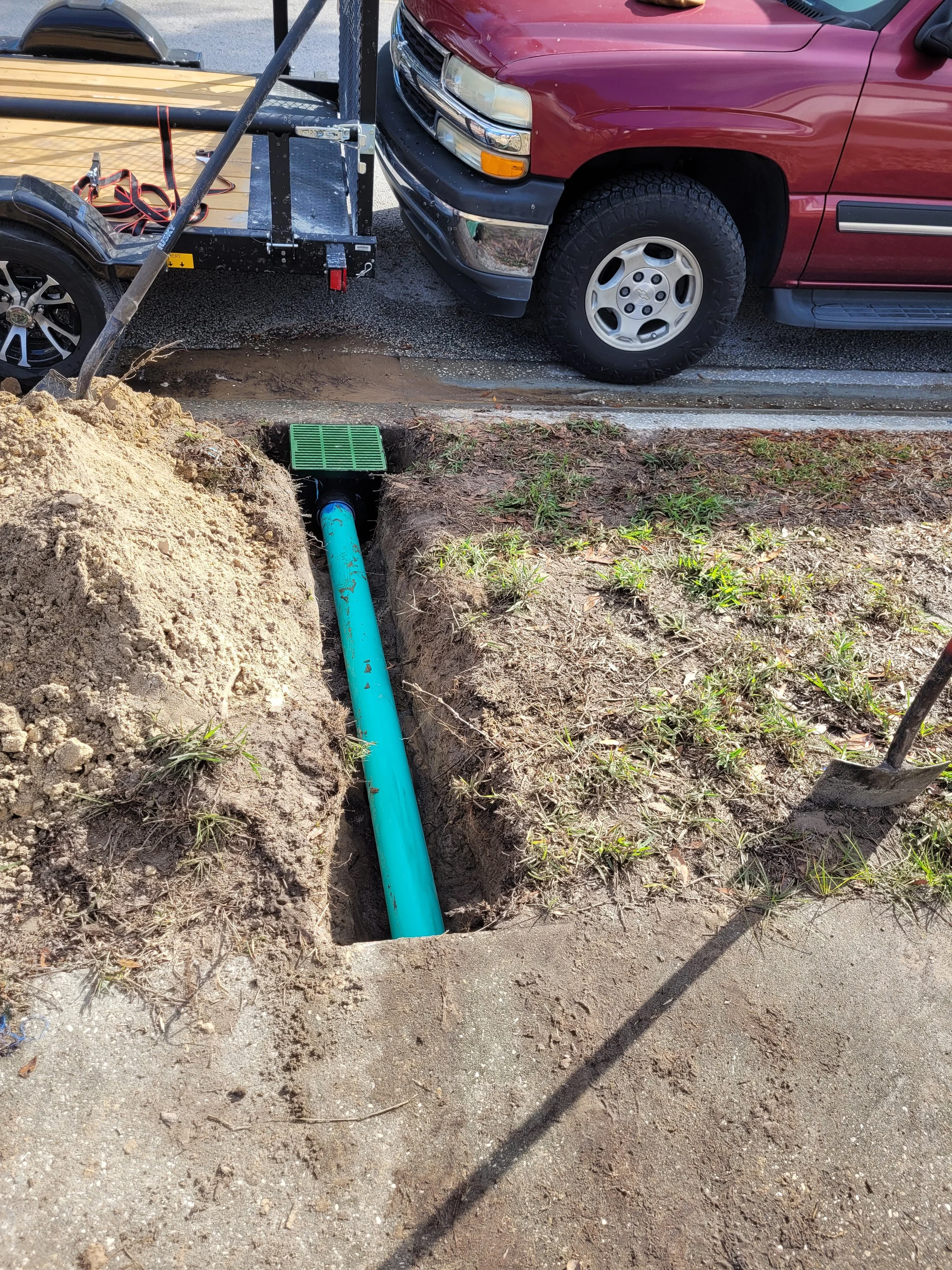 A construction site with a large orange hole in the ground, showing a blue pipe and green drainage grate. In the background, a red pickup truck is parked near a trailer with a black wheel, on a local street.