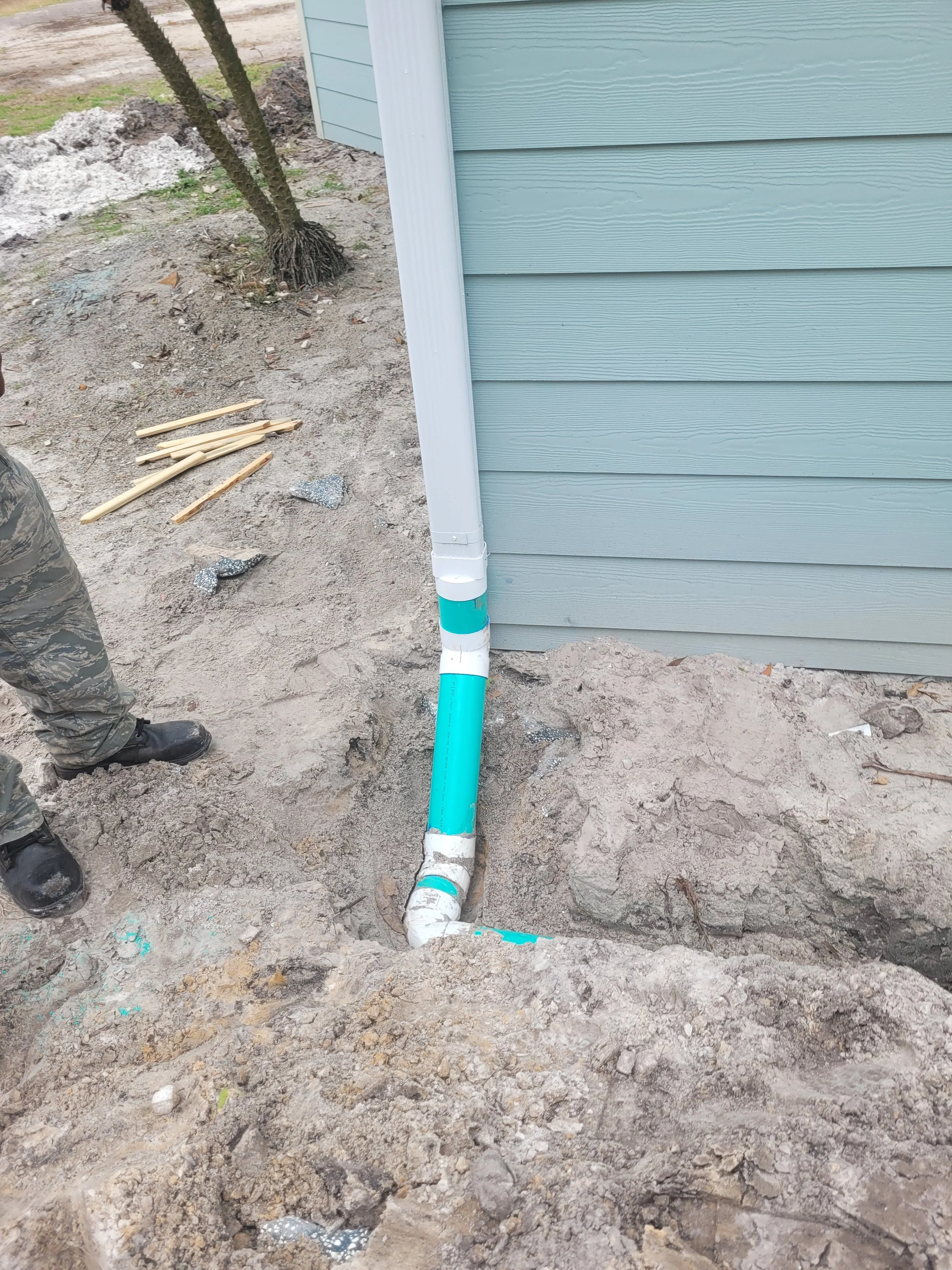 Installation of a new white and blue drain pipe next to the side of a house with light blue siding, with a trench dug in the ground for the pipe. A person's legs and boots are visible on the left side of the image.