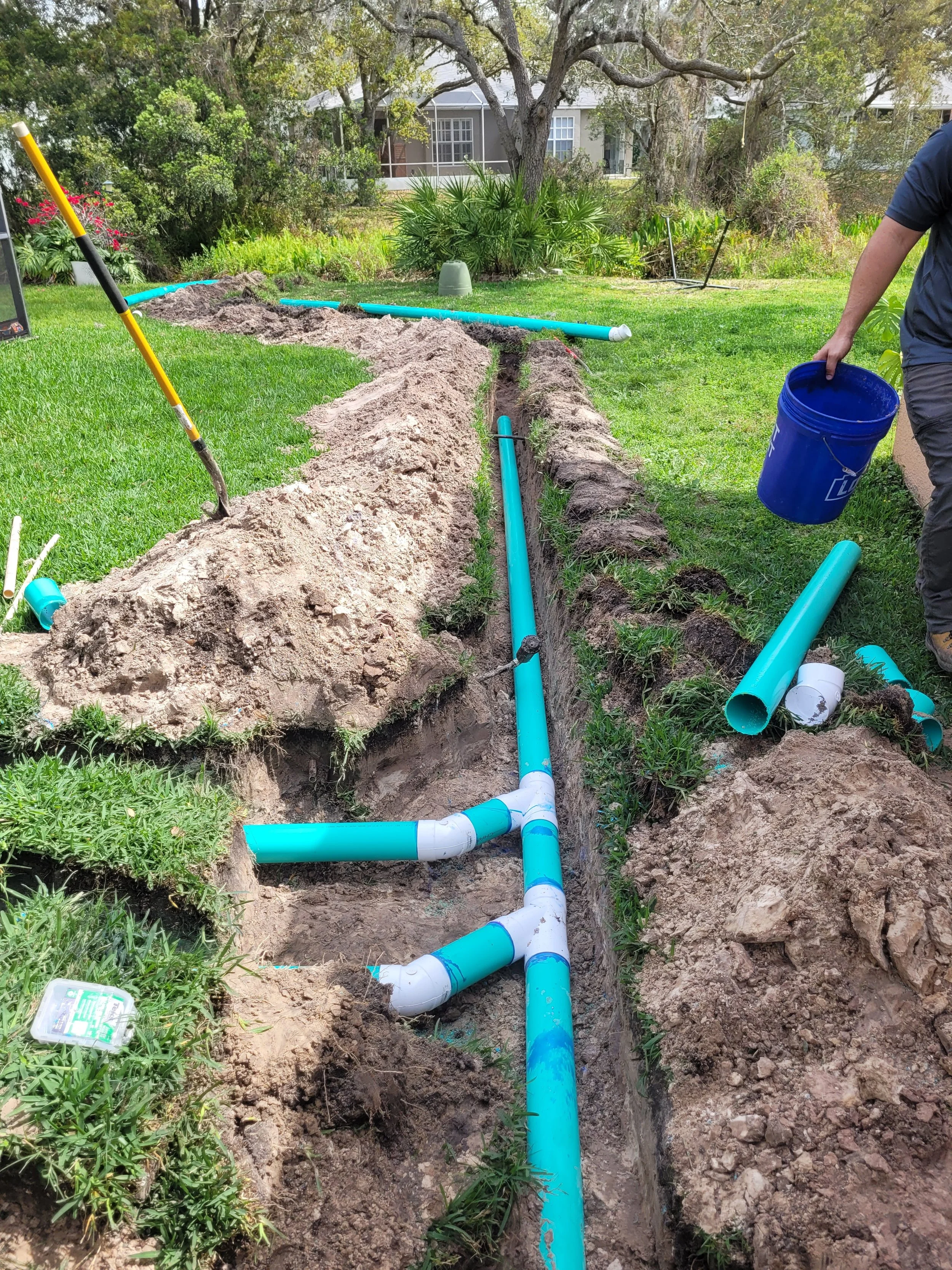 A person installing or repairing underground sprinkler irrigation pipes in a yard with green grass and trees. The pipes are blue and white, laid out in a trench dug in the ground. The person is holding a blue bucket and standing near the trench with 