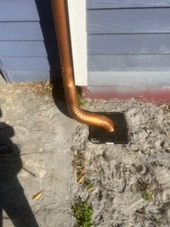 Copper downspout extending into a ground drain beside a blue house wall.