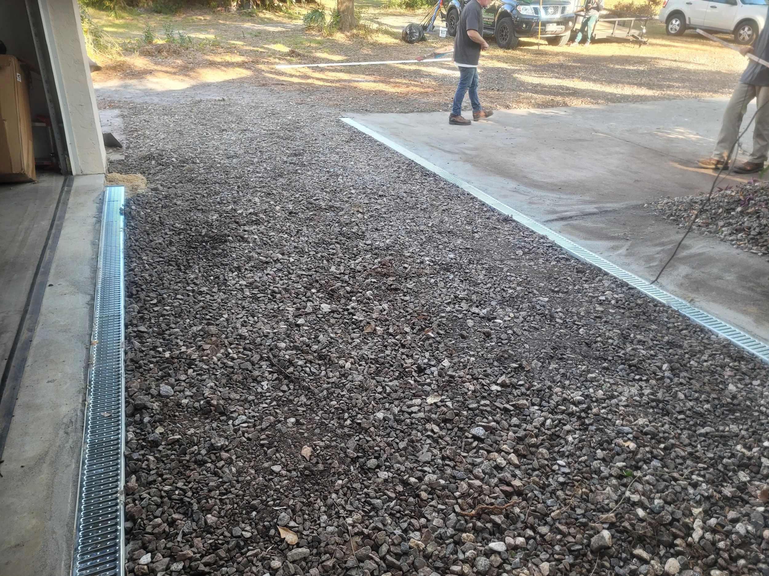 People working on a concrete driveway, using a street broom, with cars parked in the background. Gravel is spread across the driveway area.