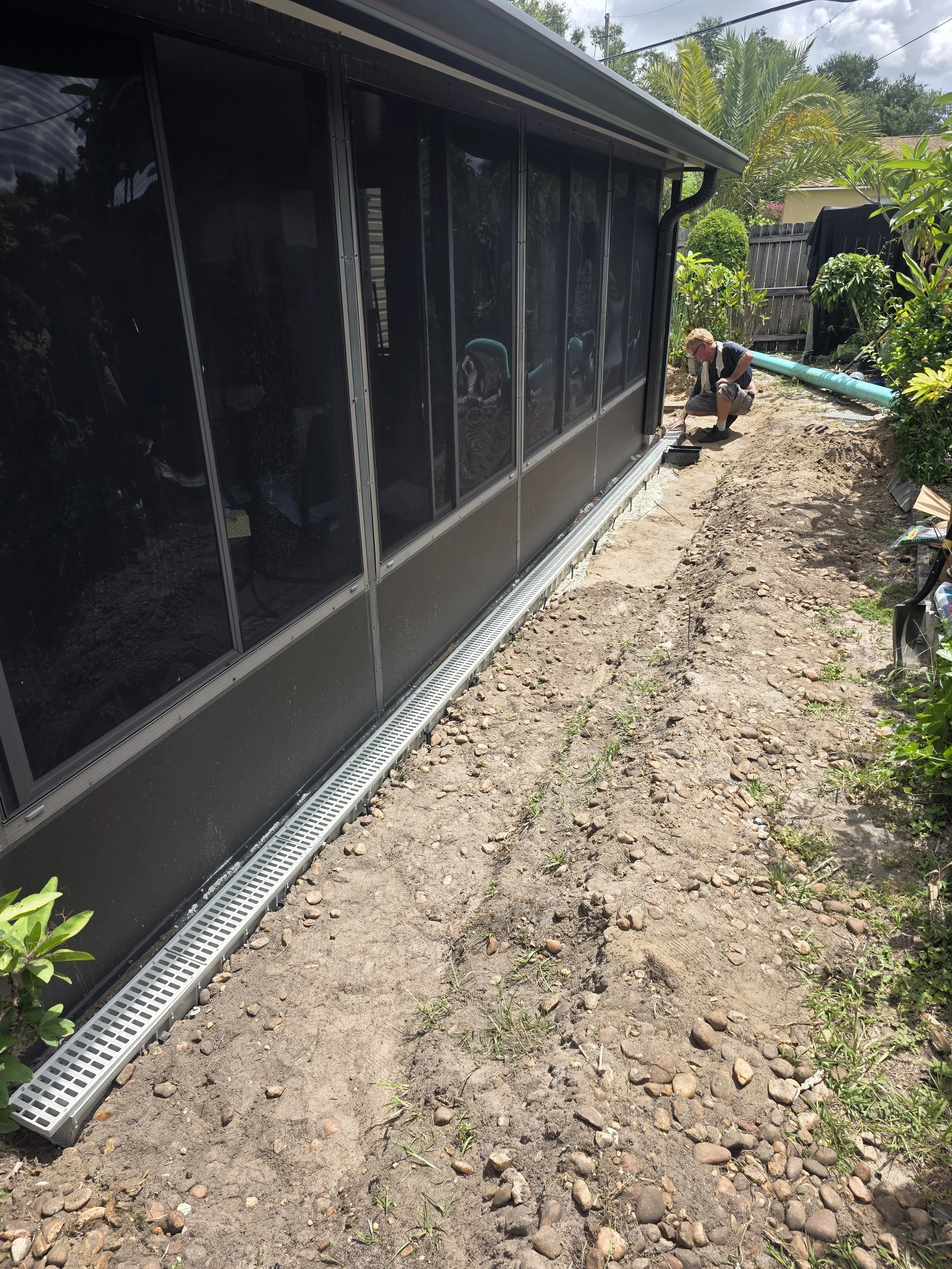 A person working outside a house with a screened porch, kneeling on the dirt near construction tools and a large blue pipe, surrounded by plants and a wooden fence.