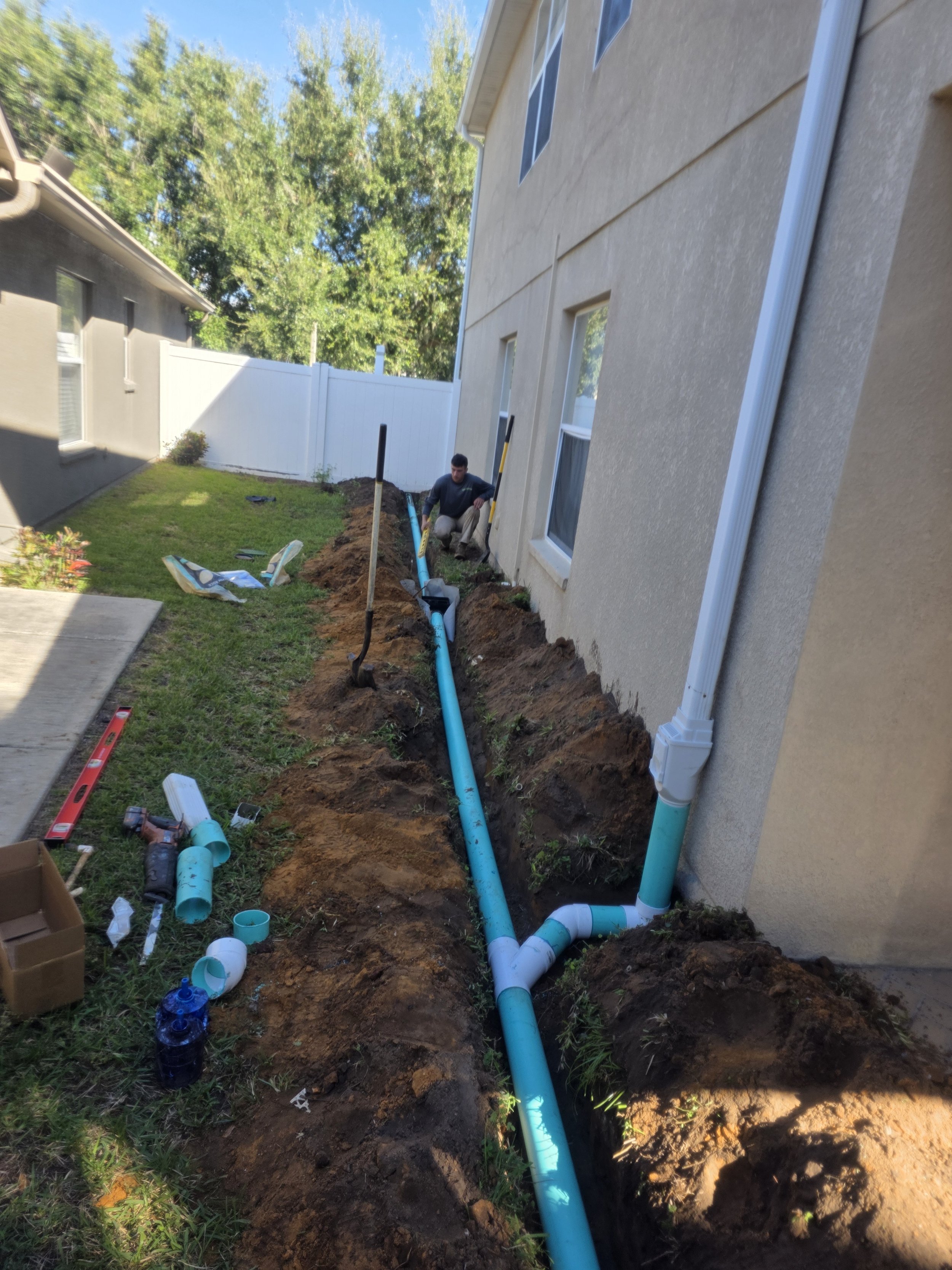 A worker installing underground drainage pipes along the side of a house in a backyard, with tools and materials nearby.