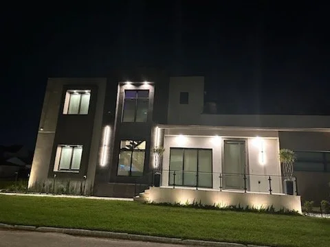 Modern two-story house at night with exterior lighting and a small balcony, surrounded by a well-manicured lawn.