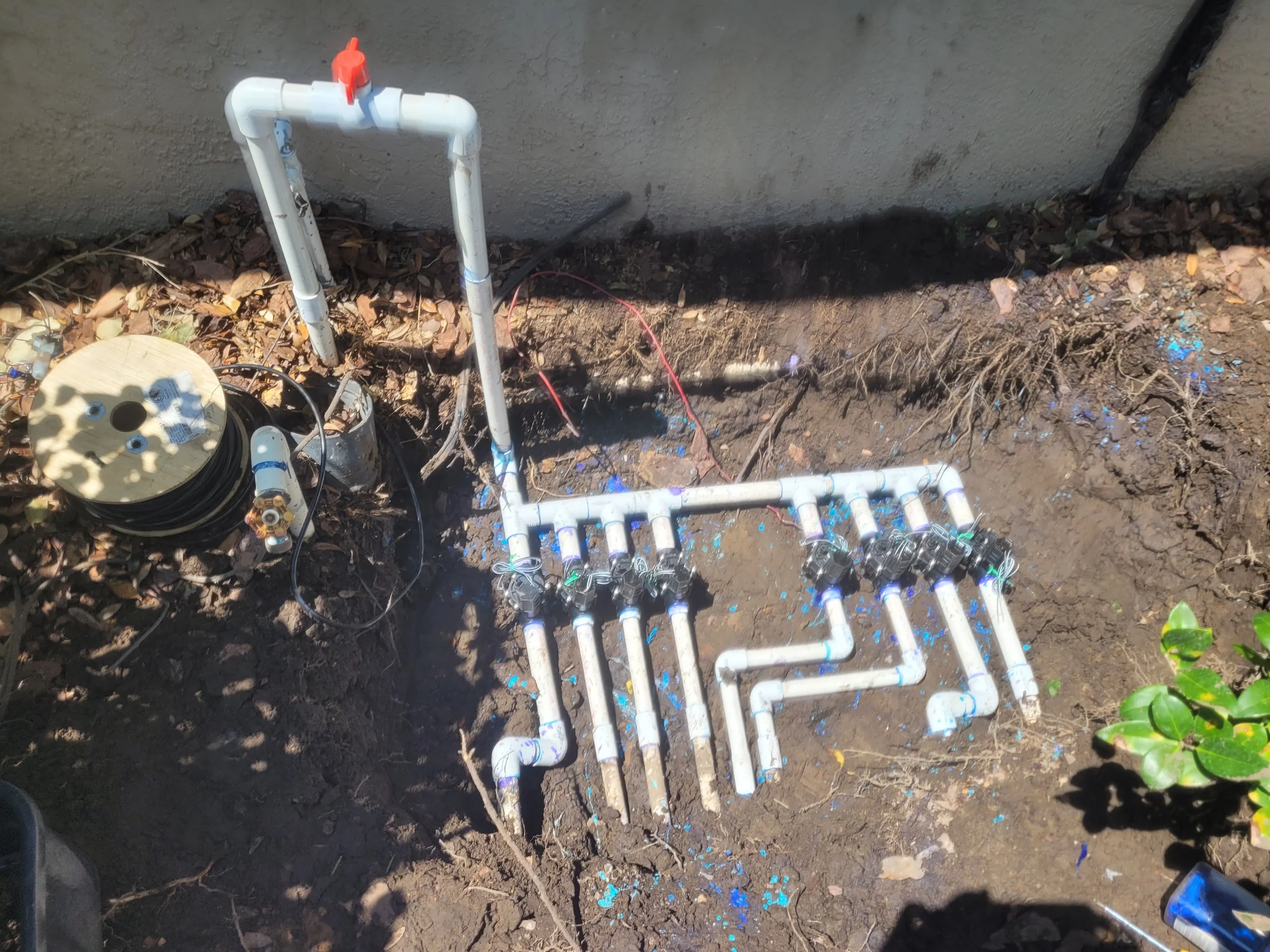 PVC pipe plumbing system installed underground with multiple pipes and valves, connected to a wall, and a reel of black tubing nearby.