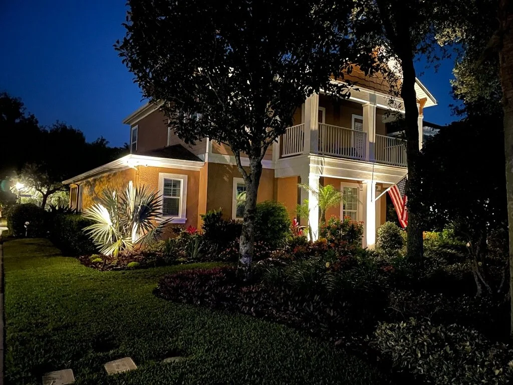 A two-story house illuminated at night, with a front porch and balcony, surrounded by lush greenery and trees, and decorated with an American flag.