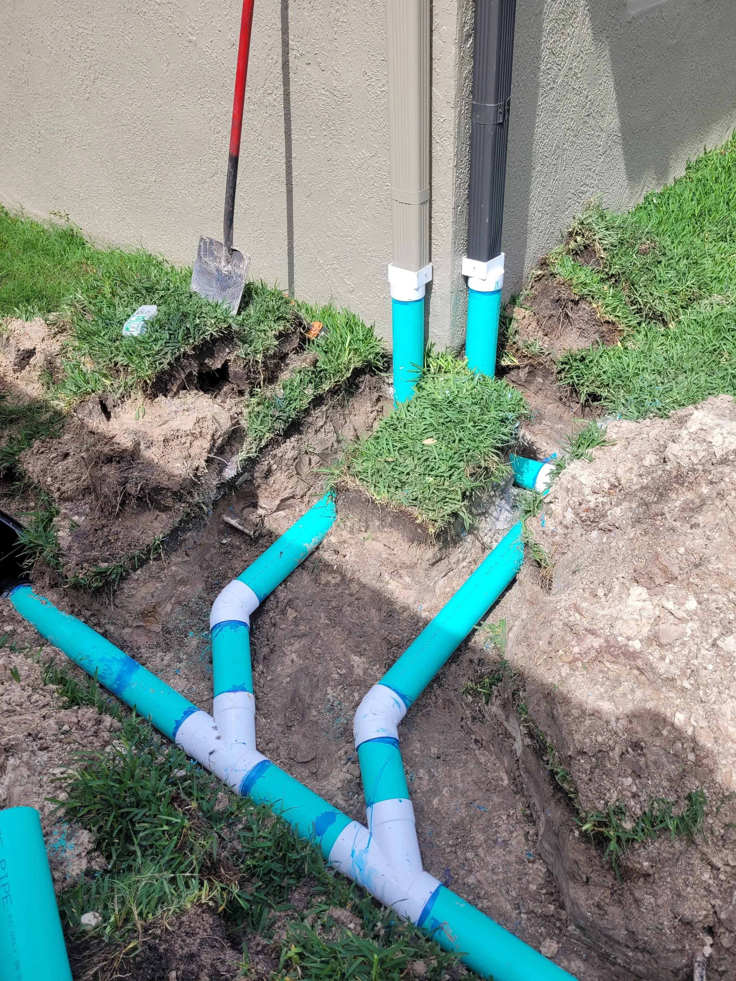 Underground utility pipes, including blue and gray pipes, are being installed outside a building near a wall with a shovel leaning against the wall. Some pipes are connected and partially buried in the soil.