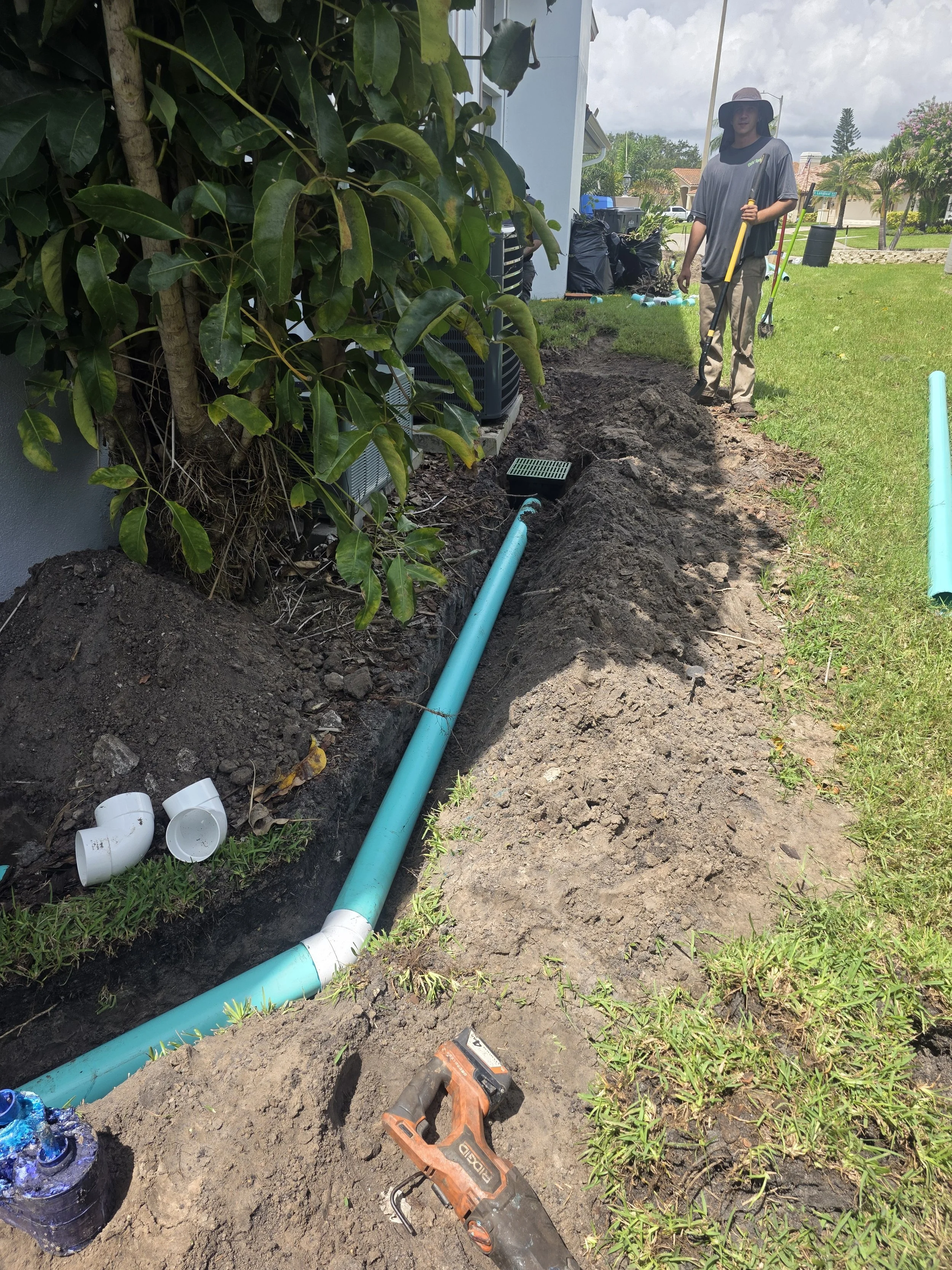 Person installing or repairing a blue PVC pipe underground next to a house, with excavation and tools visible, including a drill, on nearby soil.