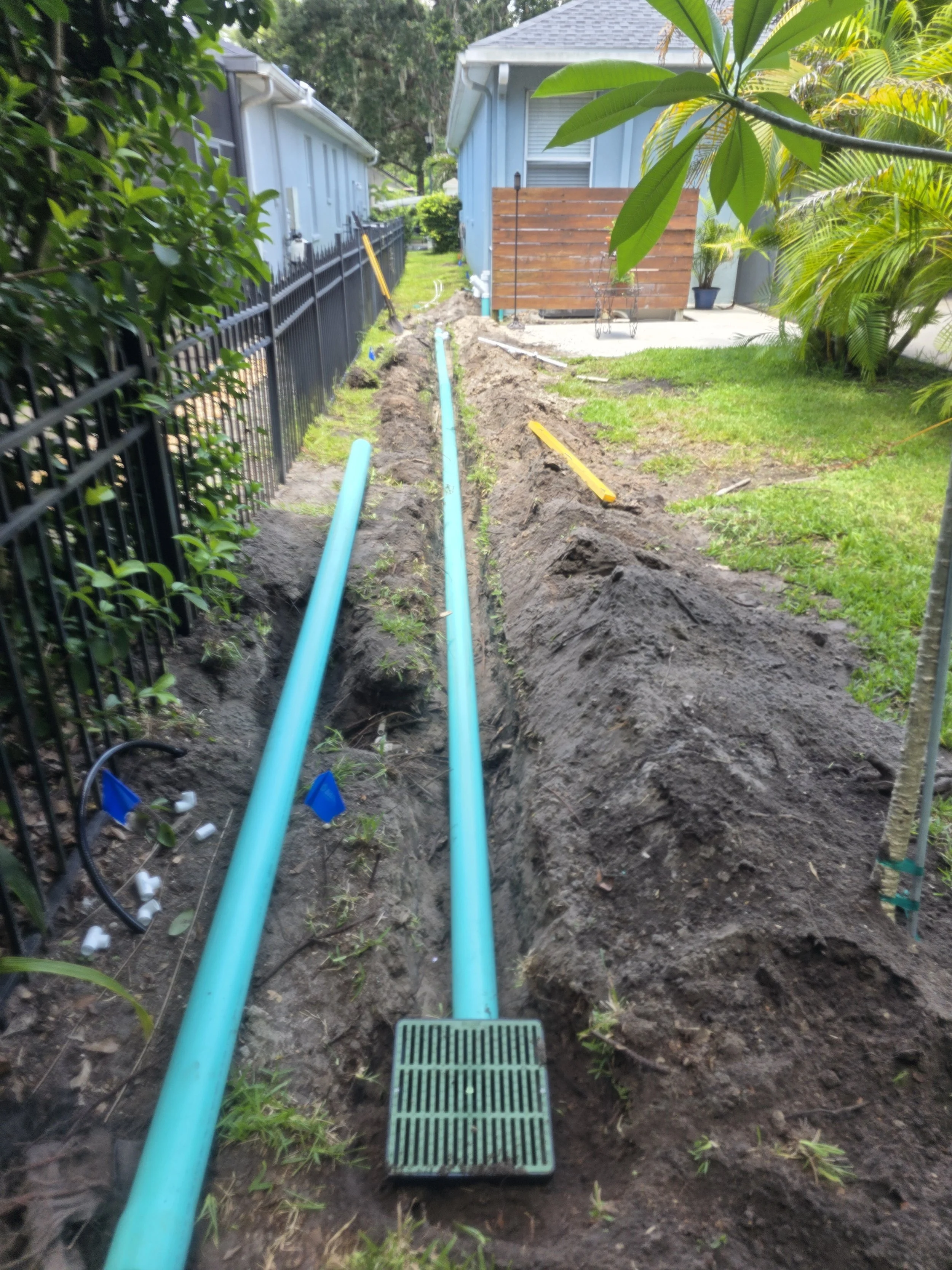 Installing or repairing underground pipes in a backyard, with exposed green pipes in a trench along a fence, next to a grassy yard and a house.