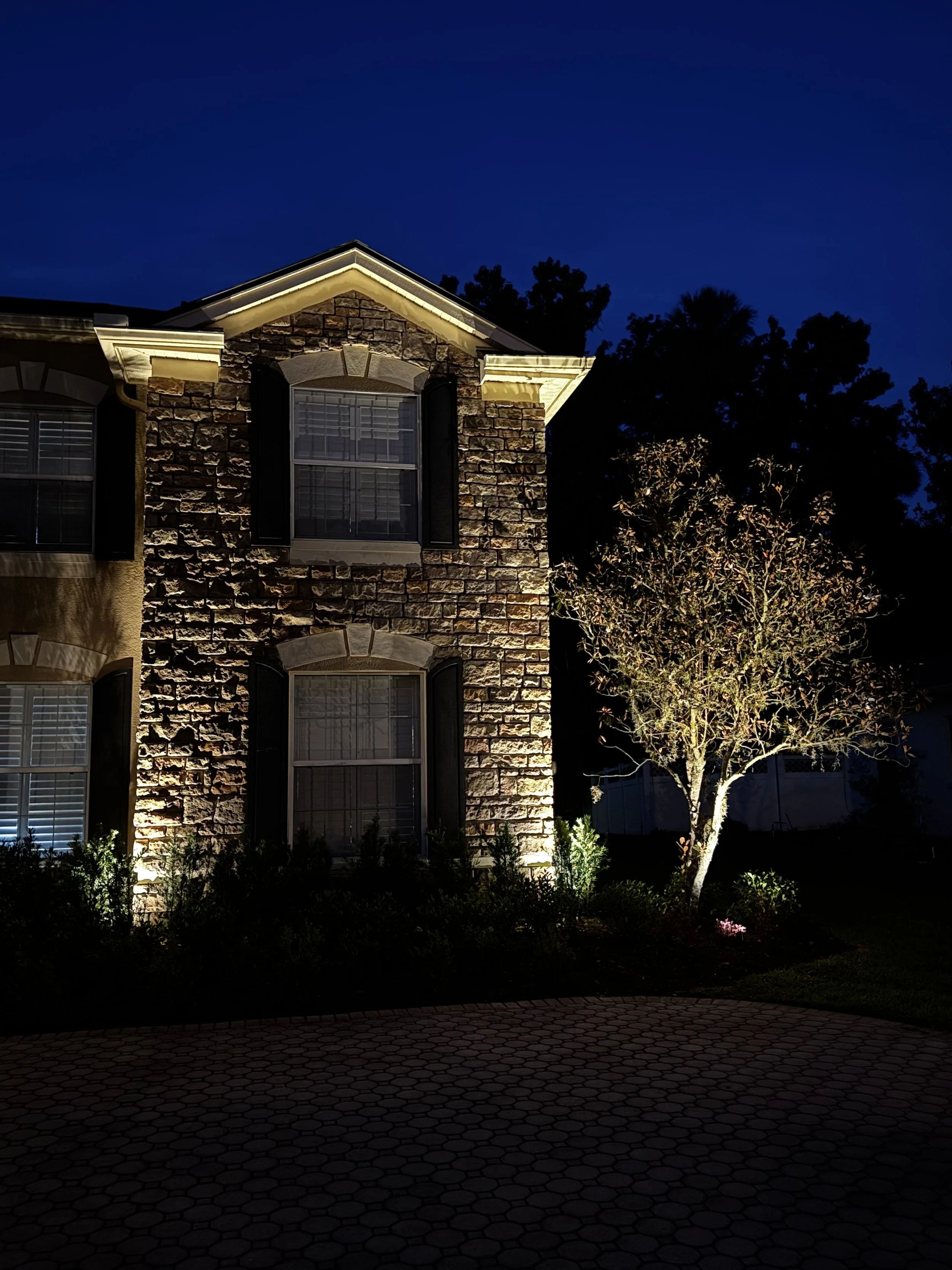 Nighttime view of a brick house with illuminated windows and exterior lighting, a tree to the right, and a brick driveway in the foreground.