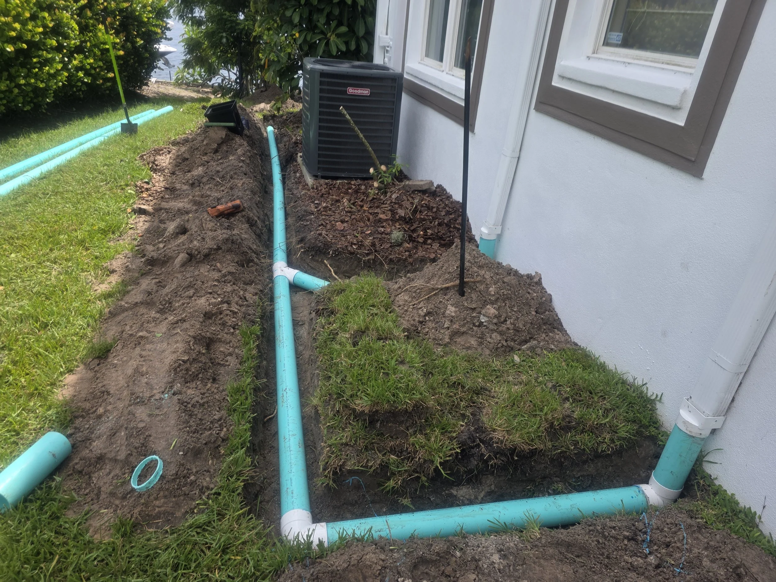Garden landscape with a trench dug for plumbing installation, including blue PVC pipes, near the side of a white house with windows, surrounded by grass, bushes, and a backyard area.