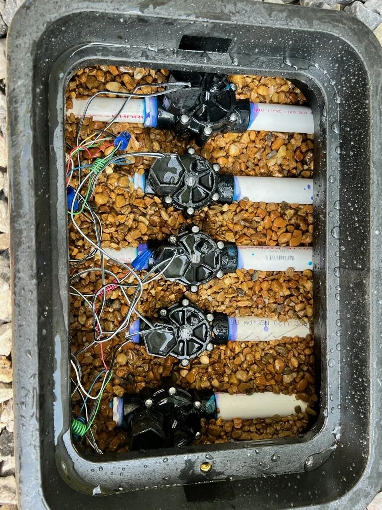 Inside a water timer box with multiple black valve components, white PVC pipes, and wiring, all set on a bed of small rocks.