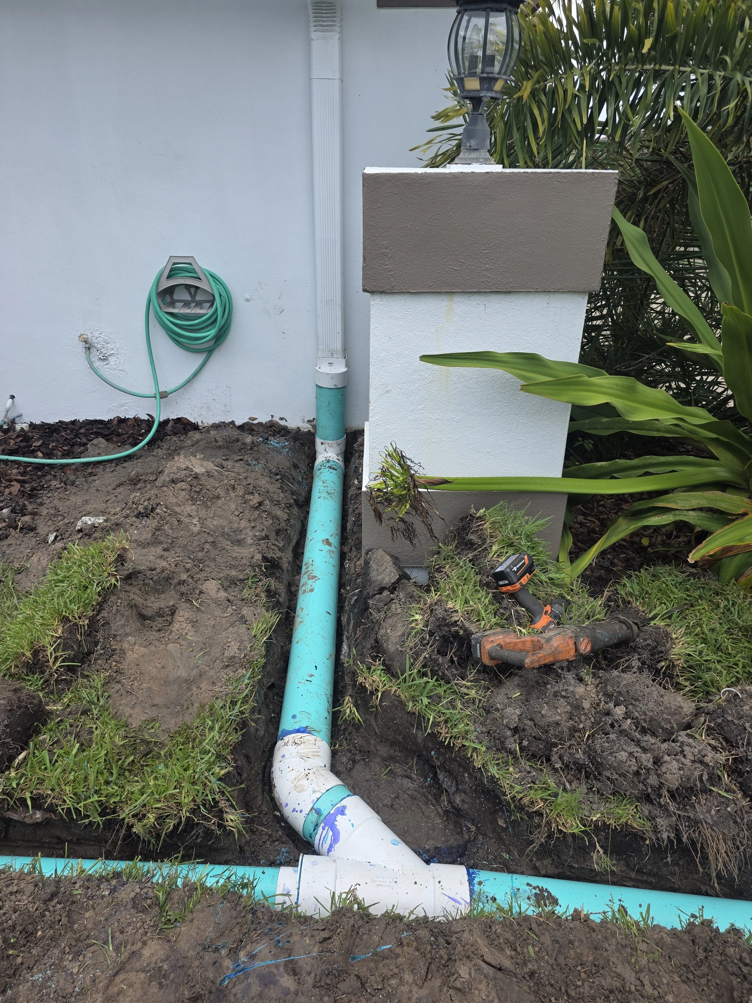 Outdoor plumbing work with exposed blue and white pipes, a cordless drill, and a black and orange screwdriver on the ground beside the foundation of a house, with a garden hose on the wall and landscaping plants.