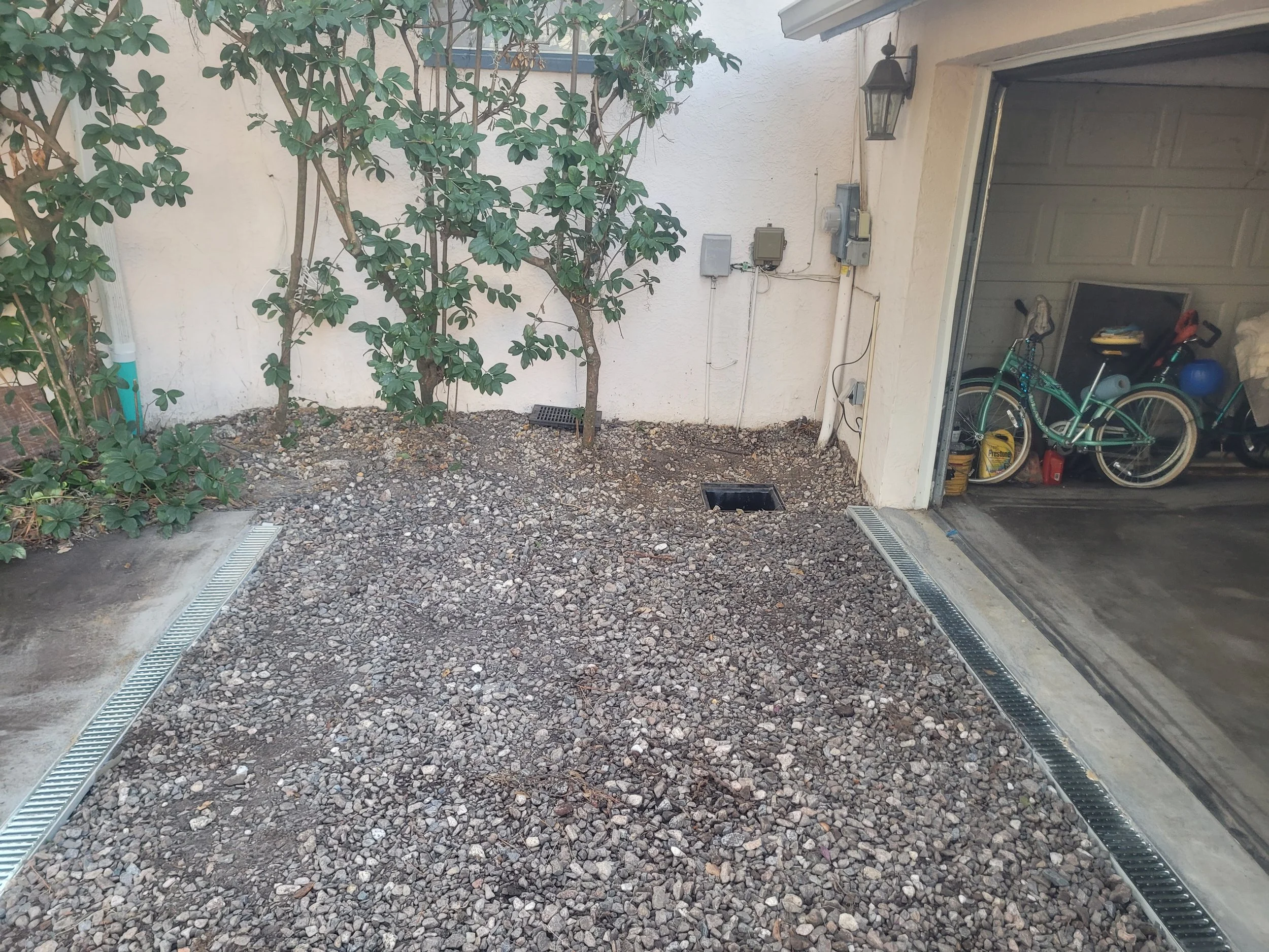 Part of a garage and backyard with gravel ground, some plants, and electrical meters on the wall.