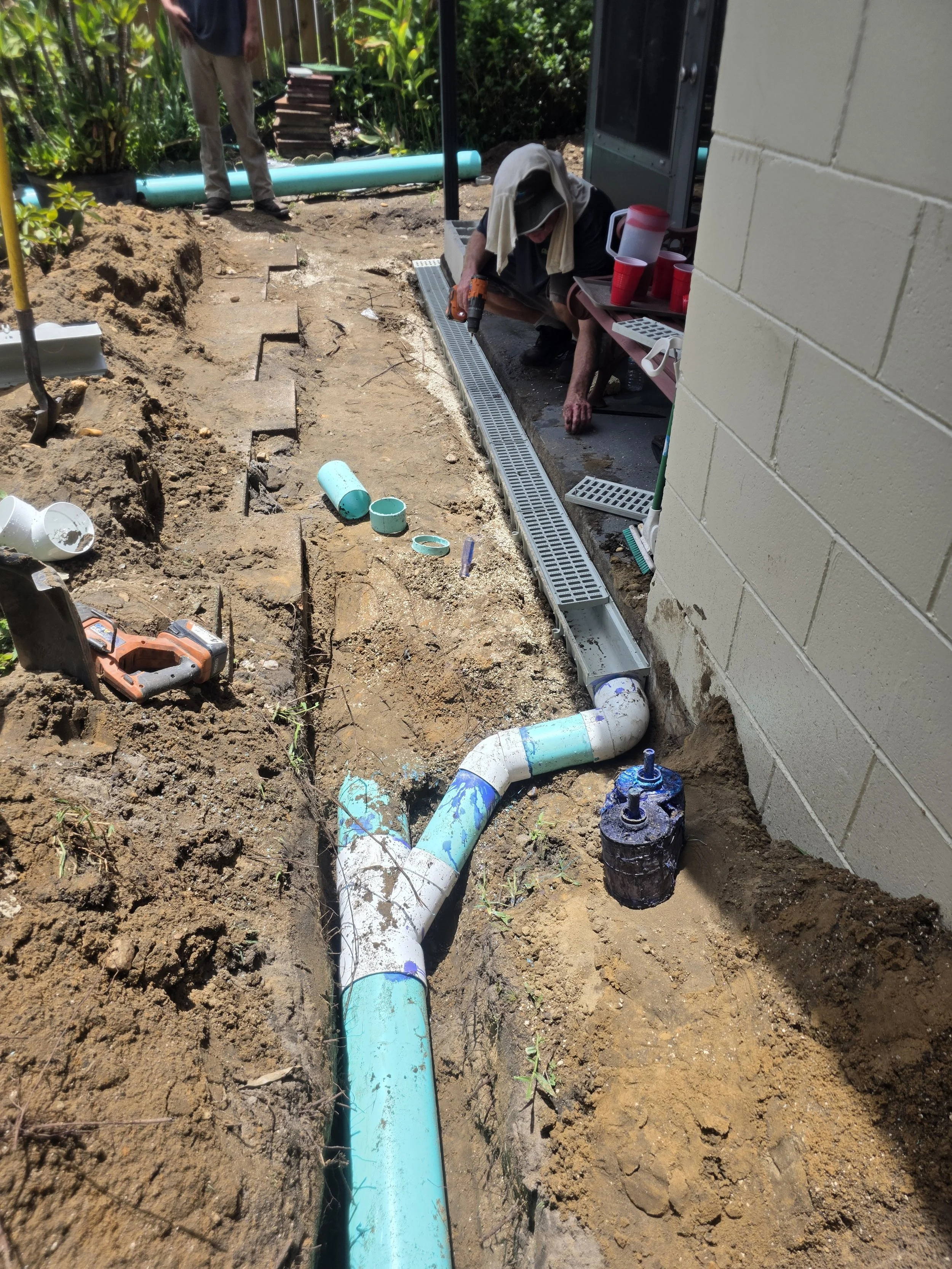 Workers installing a drainage system outside a building, with one worker crouching and another standing nearby, surrounded by construction tools and pipes.