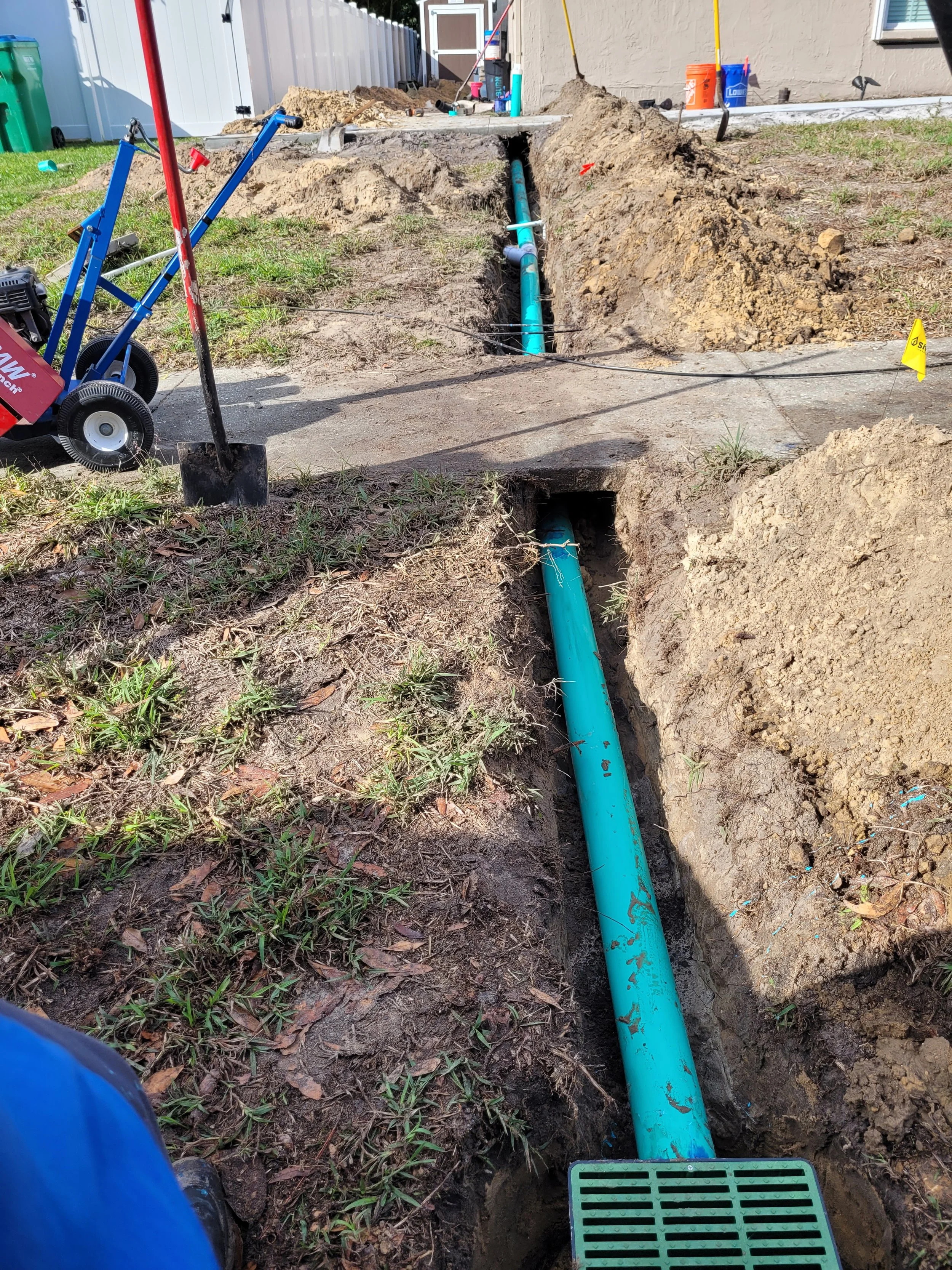 Construction site with a trench in the ground containing a blue pipe, surrounded by dirt and grass, with tools and construction materials visible in the background.