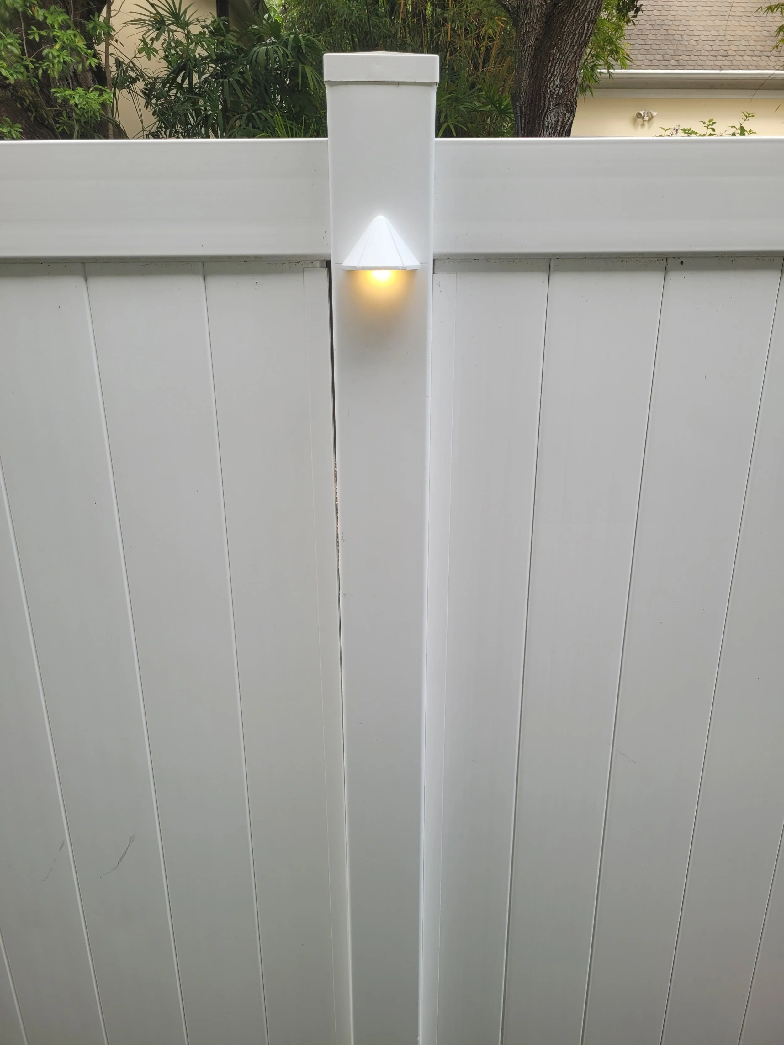 A white outdoor wall sconce light fixture mounted on a white fence, illuminating with a yellowish glow.