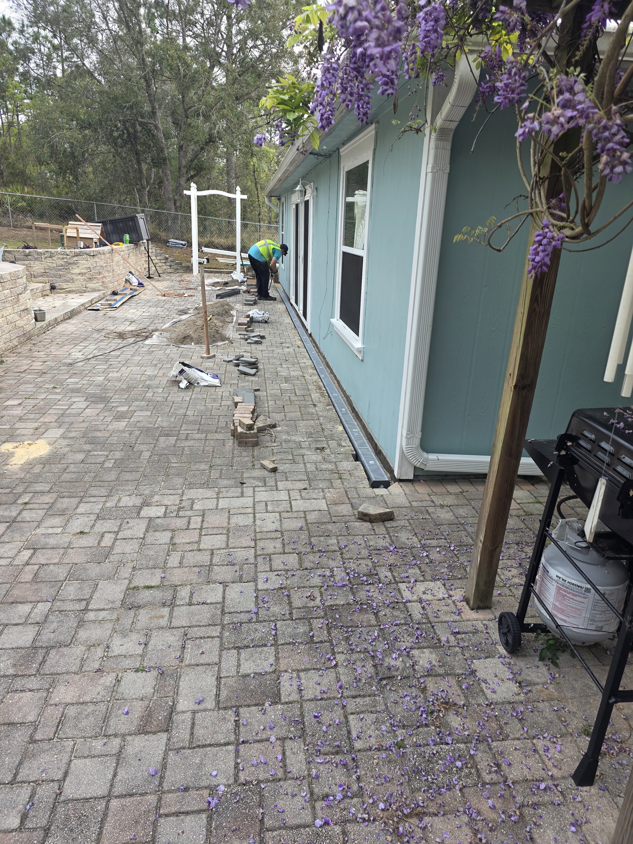 Person working on installing or repairing a sliding glass door on a house exterior with a brick patio, purple flowers hanging from a tree, and construction tools and materials scattered around.