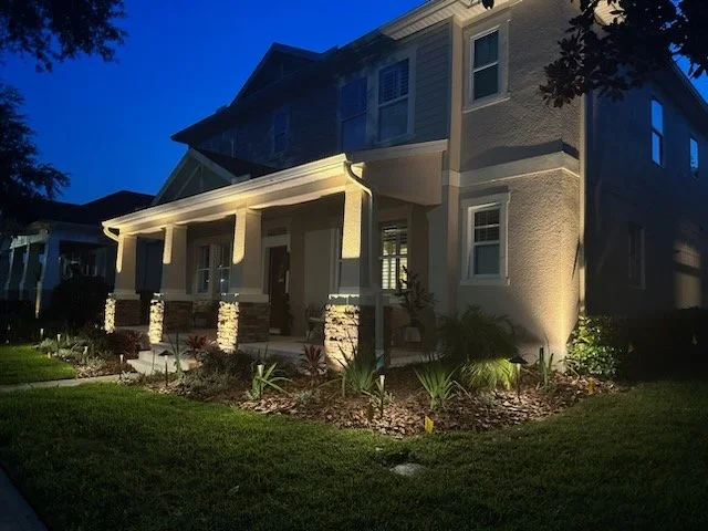 A well-lit house at dusk with a front porch, stone columns, and landscaped garden in the foreground.