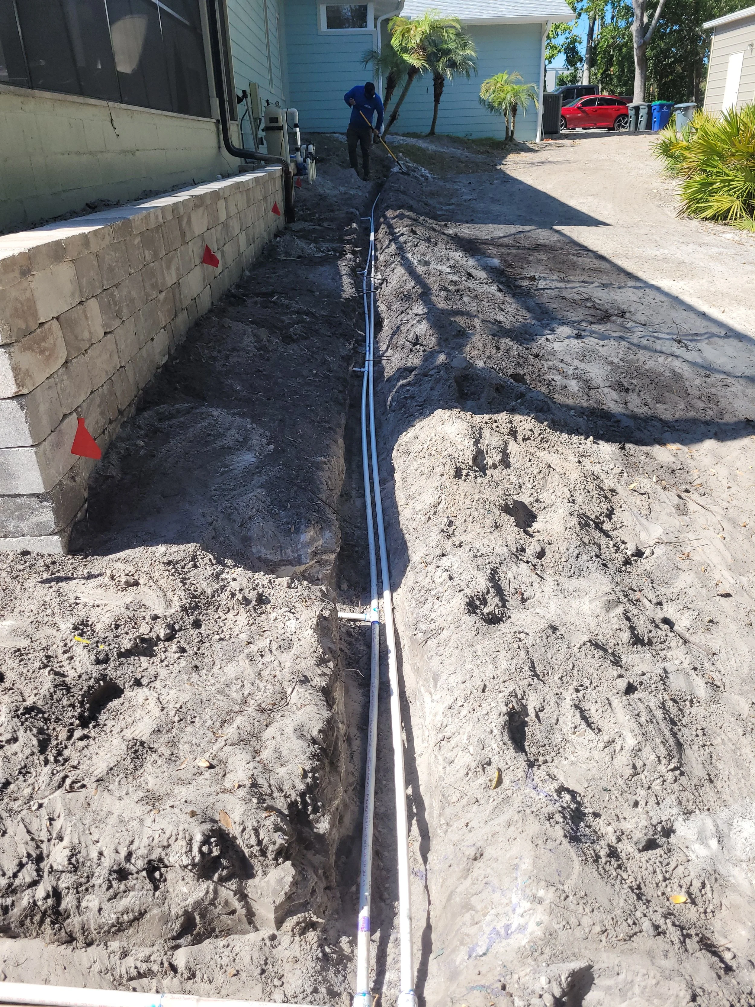 Construction worker installing underground pipes next to a house, with a retaining wall on the side and a dirt pathway.