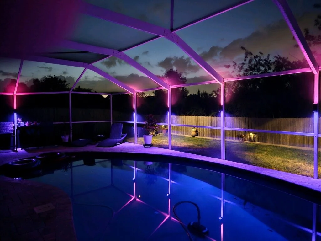 An enclosed backyard pool area at dusk with purple and pink LED lighting along the fence and ceiling framework, with outdoor furniture and plants visible.