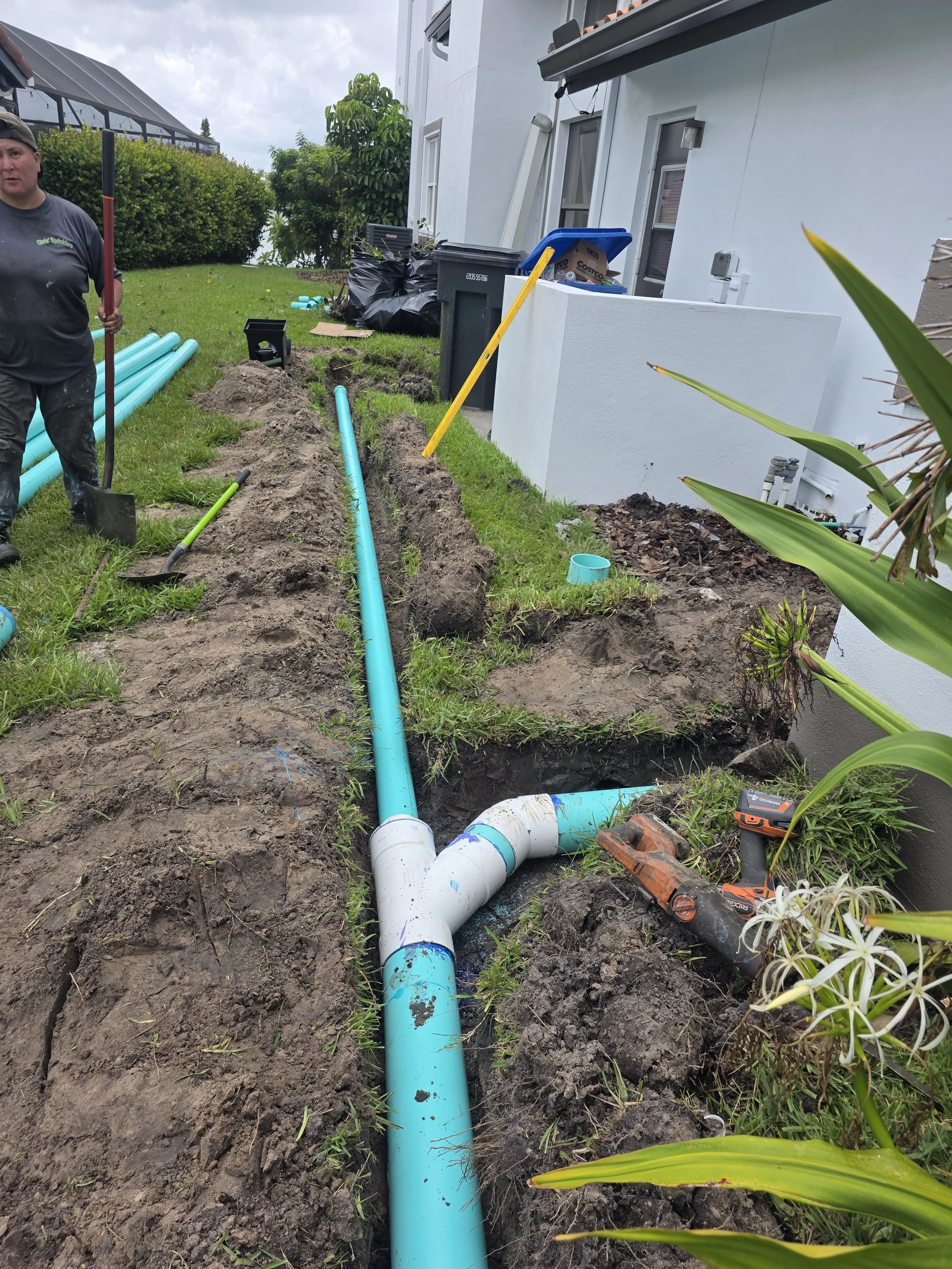Workers installing blue PVC pipes in a trench outside a white building with a lawn and plants nearby.
