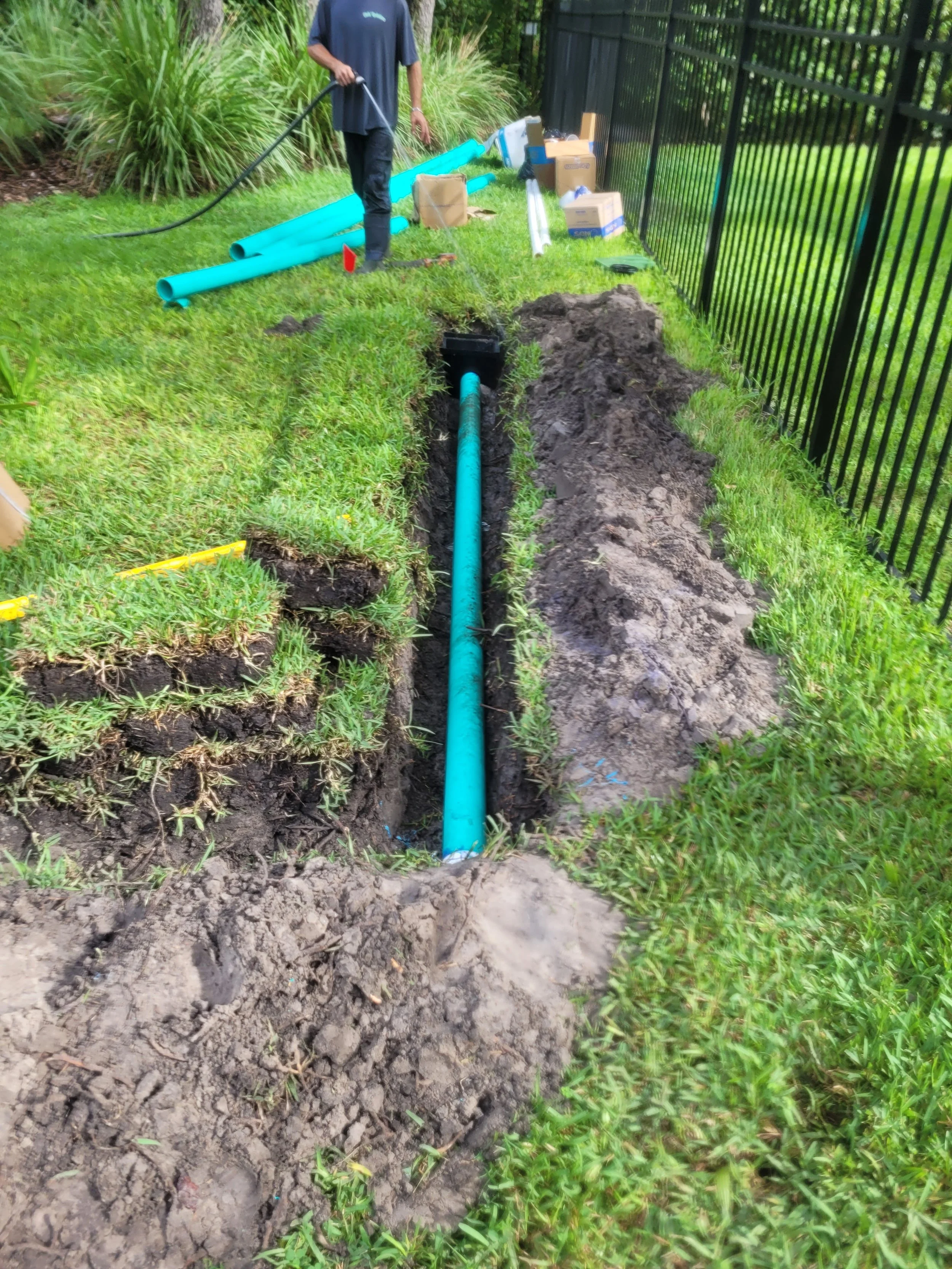 Inspection or installation of a blue underground pipe in a grassy outdoor area next to a black metal fence.
