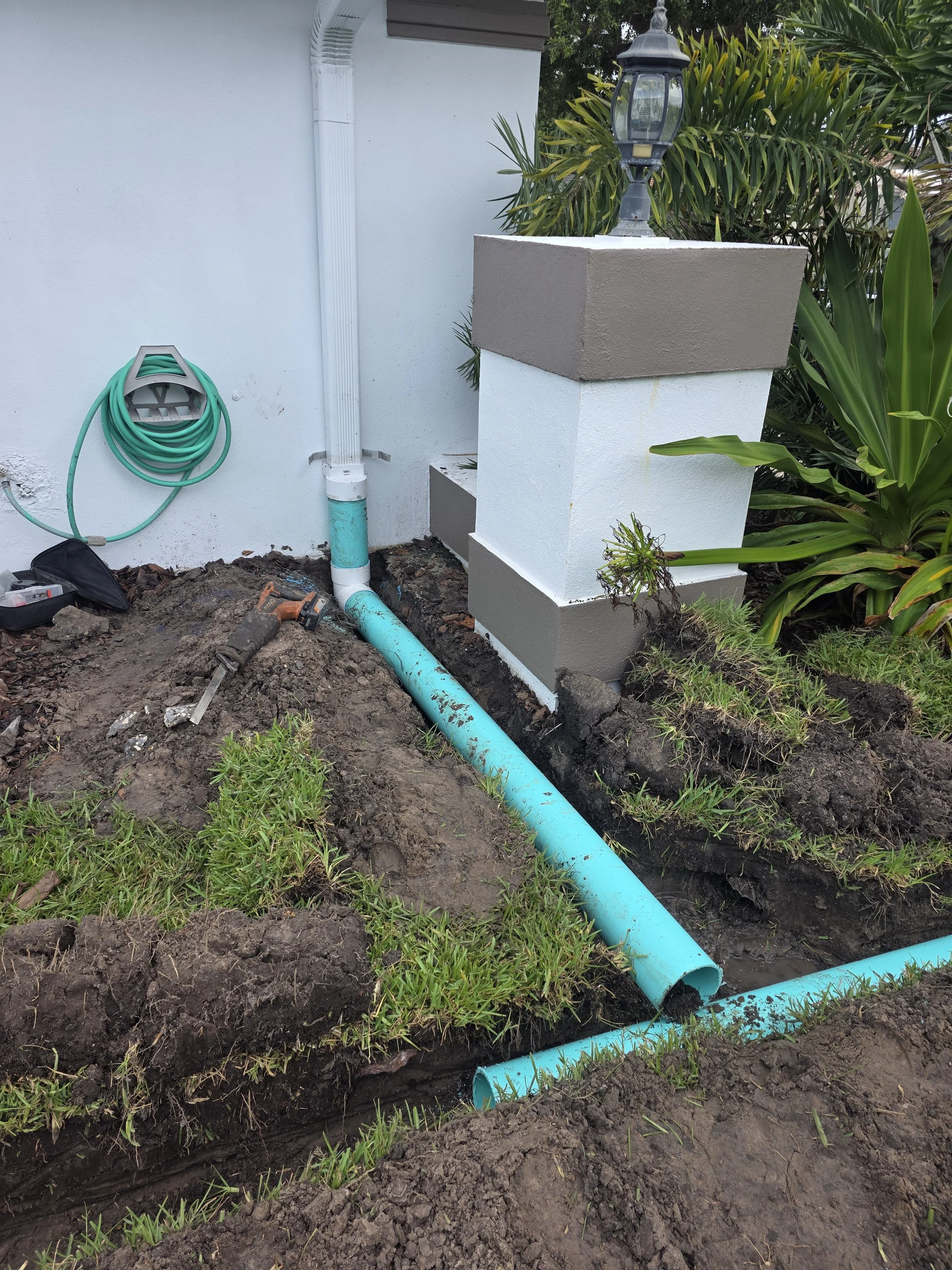 Underground plumbing installation with blue pipes, a drainage pipe, a drain snake, and tools near a house wall with a white and brown outdoor light fixture and green plants.
