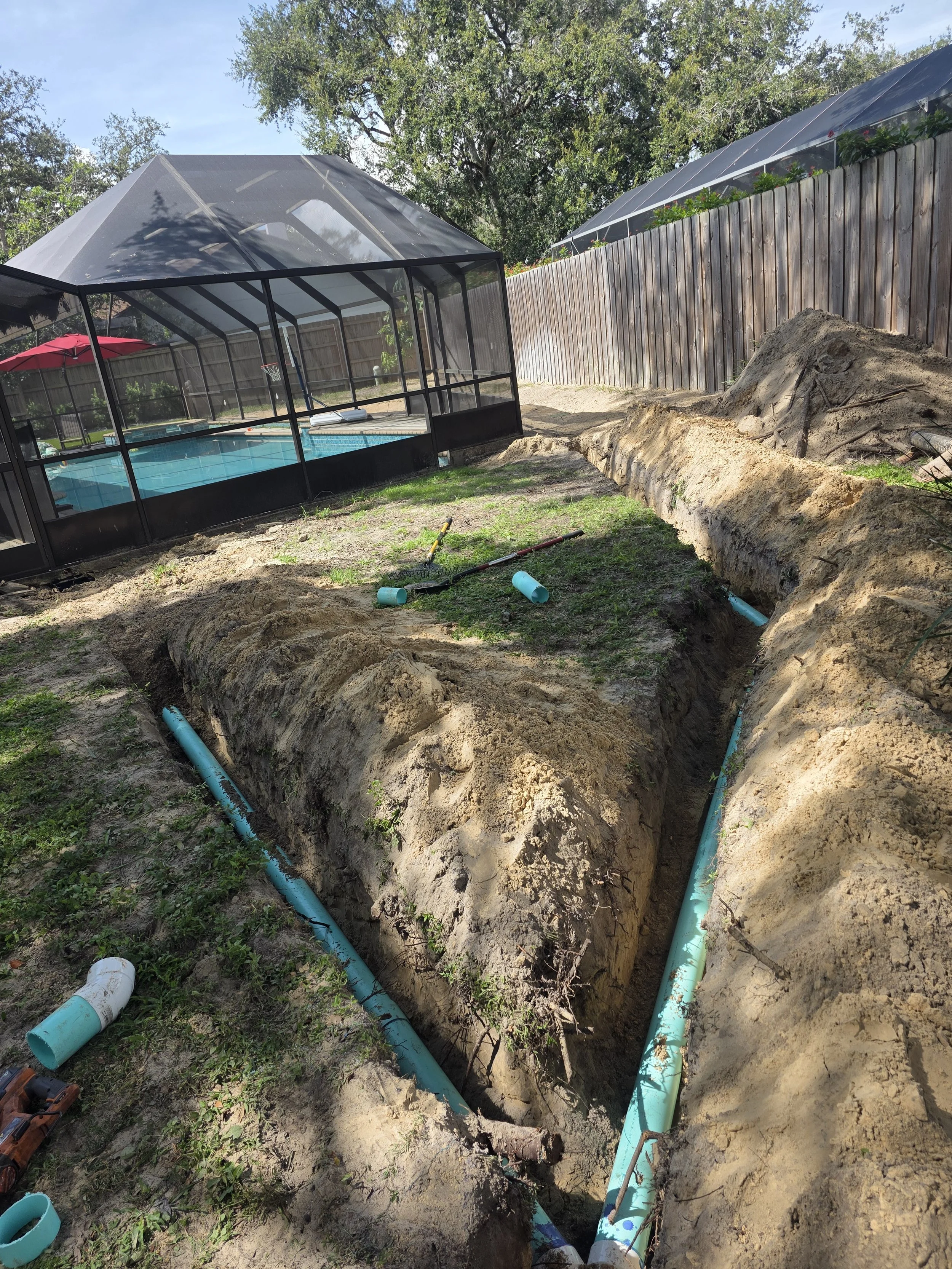 Digging trench for underground pipe installation next to a backyard, with a screened pool enclosure, a wooden fence, and trees in the background.