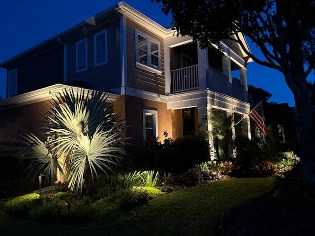 A two-story house at night with exterior lighting, a front porch with an American flag, and landscaped yard with a large plant and trees.