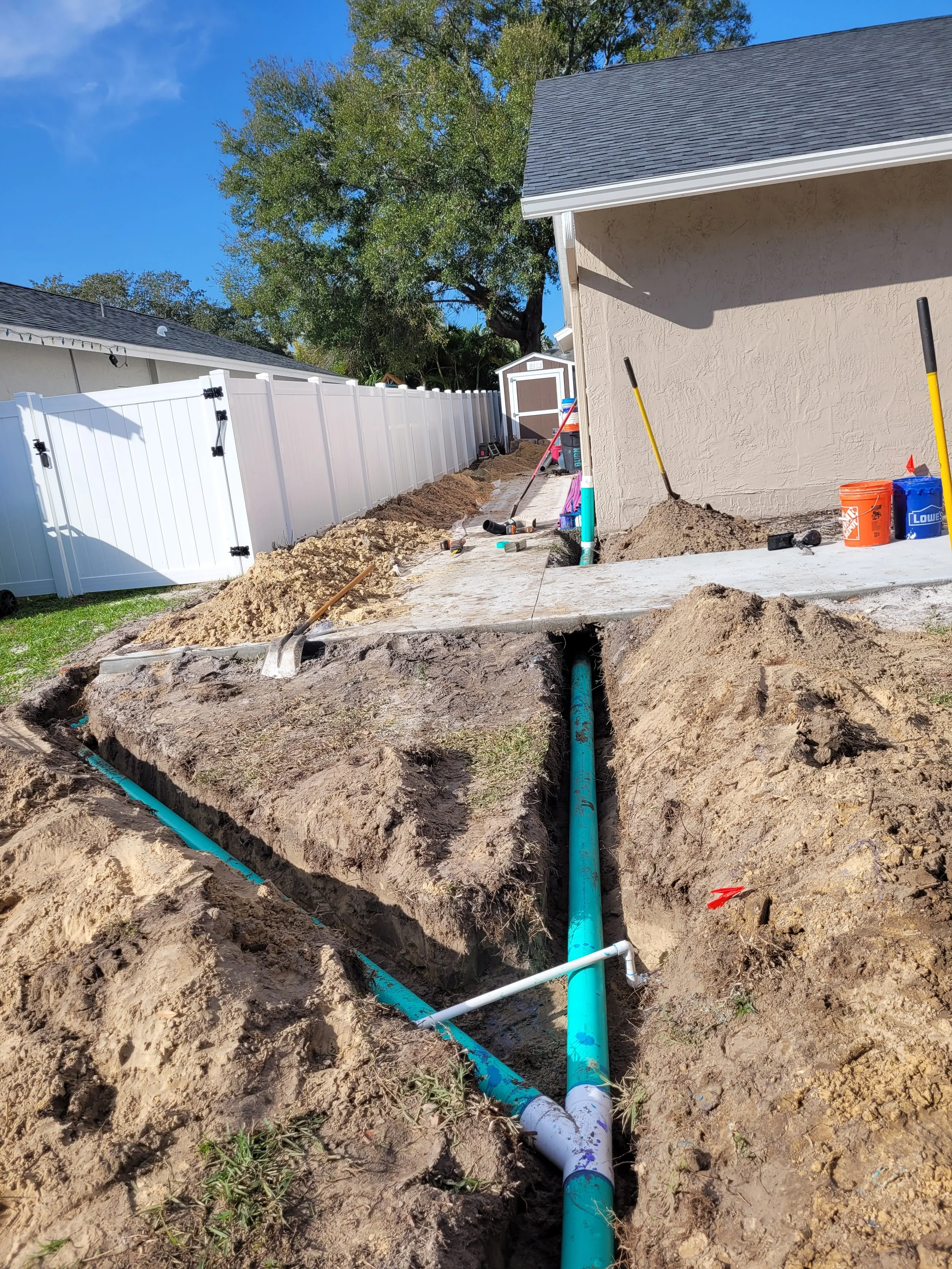Underground plumbing work around a house with exposed pipes, tools, and construction materials in a backyard.