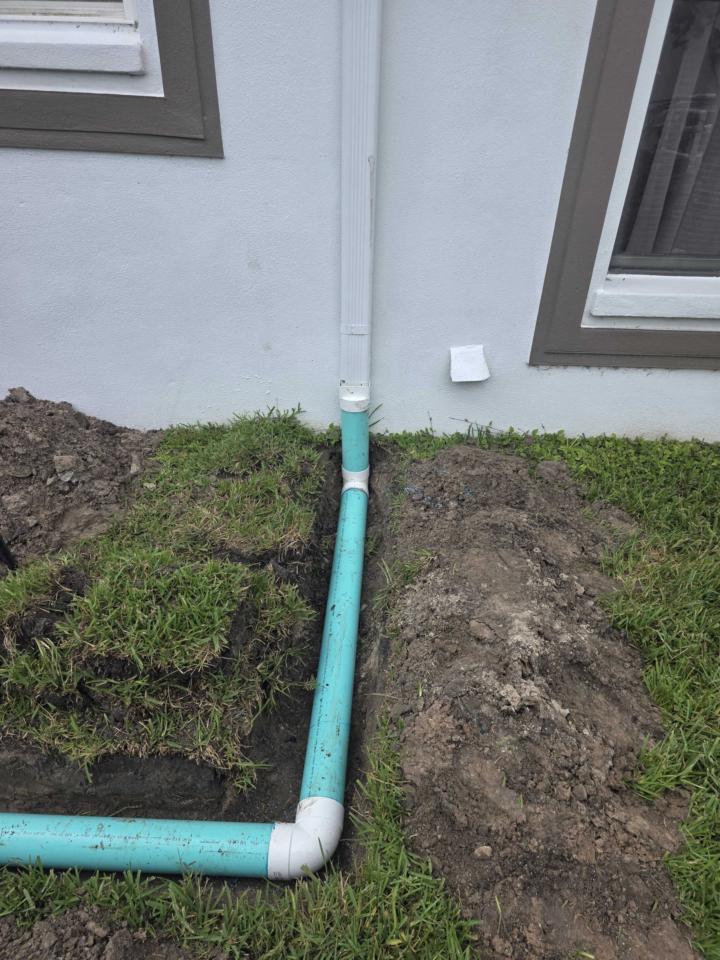 Underground plumbing pipe installation next to a house exterior wall with two brown and white framed windows.