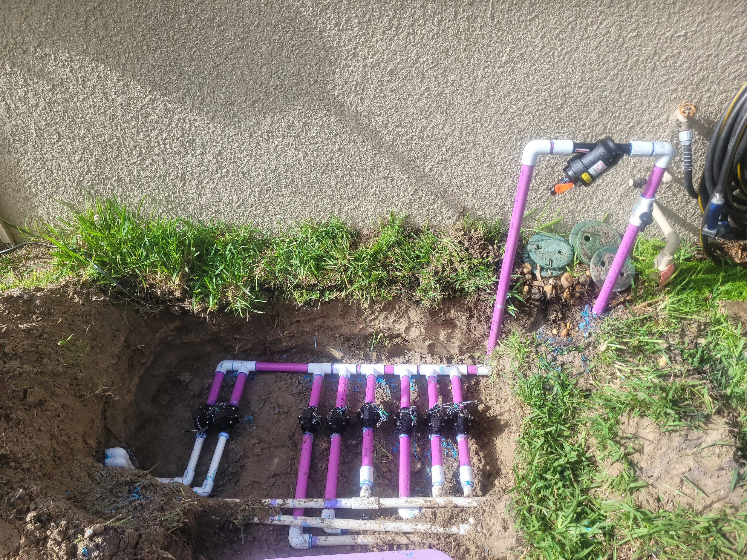 An irrigation system with connected pipes in a shallow trench next to a beige wall, with grass and zucchini plants nearby.